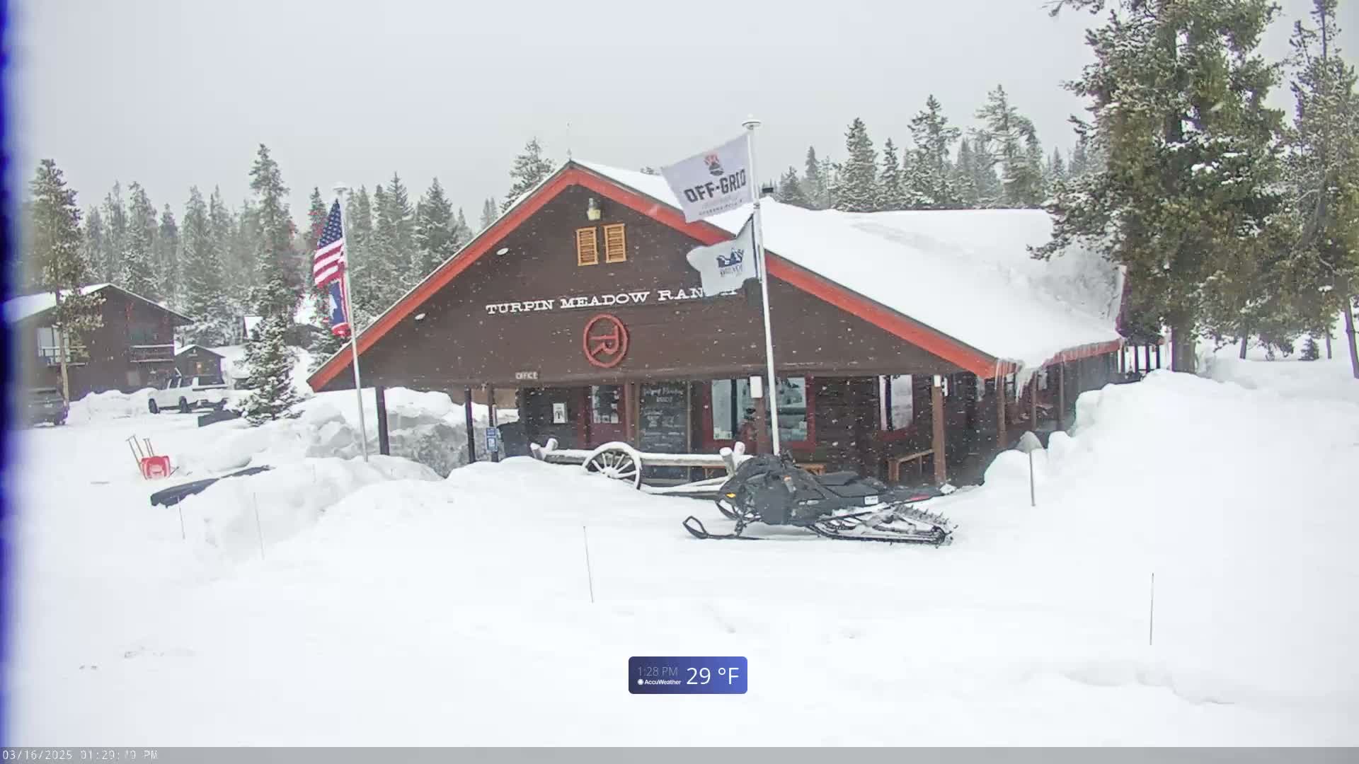 A snow-covered building, possibly a lodge or ranch, sits in a snowy landscape under a light snow, with a snowmobile parked nearby.