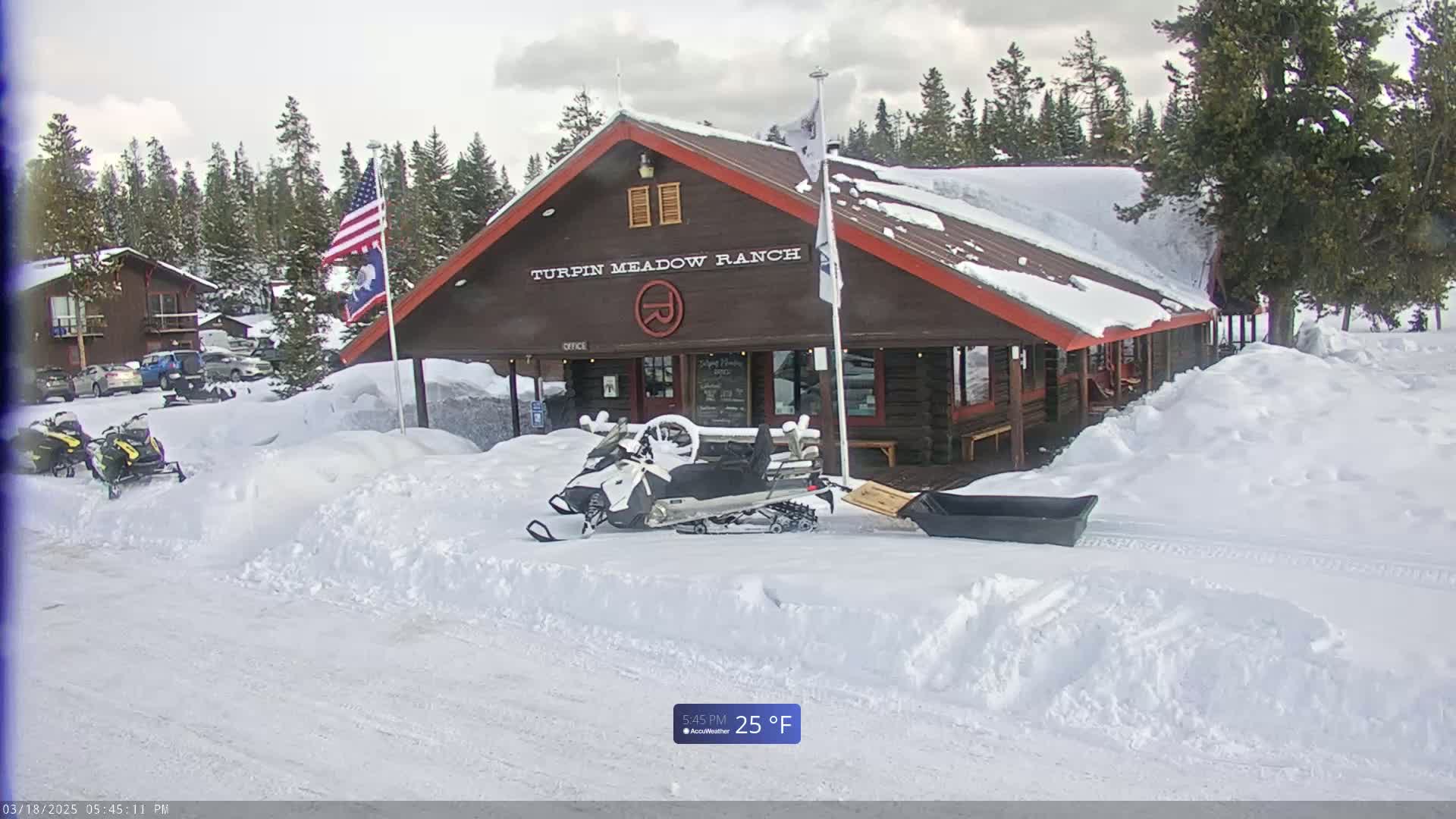 A snow-covered log cabin building, with snowmobiles parked in front, sits in a snowy landscape under a cloudy sky.