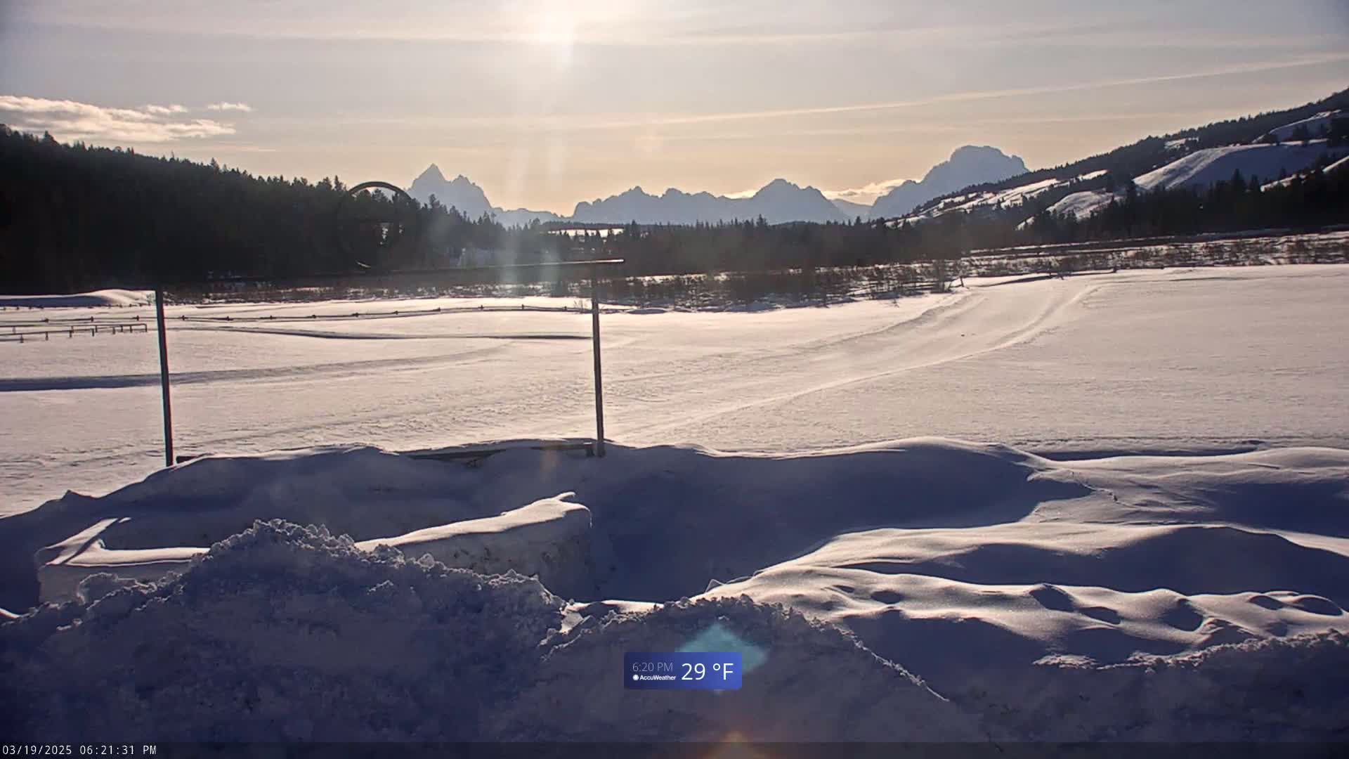 A snow-covered field stretches out before a line of mountains under a sunny sky.