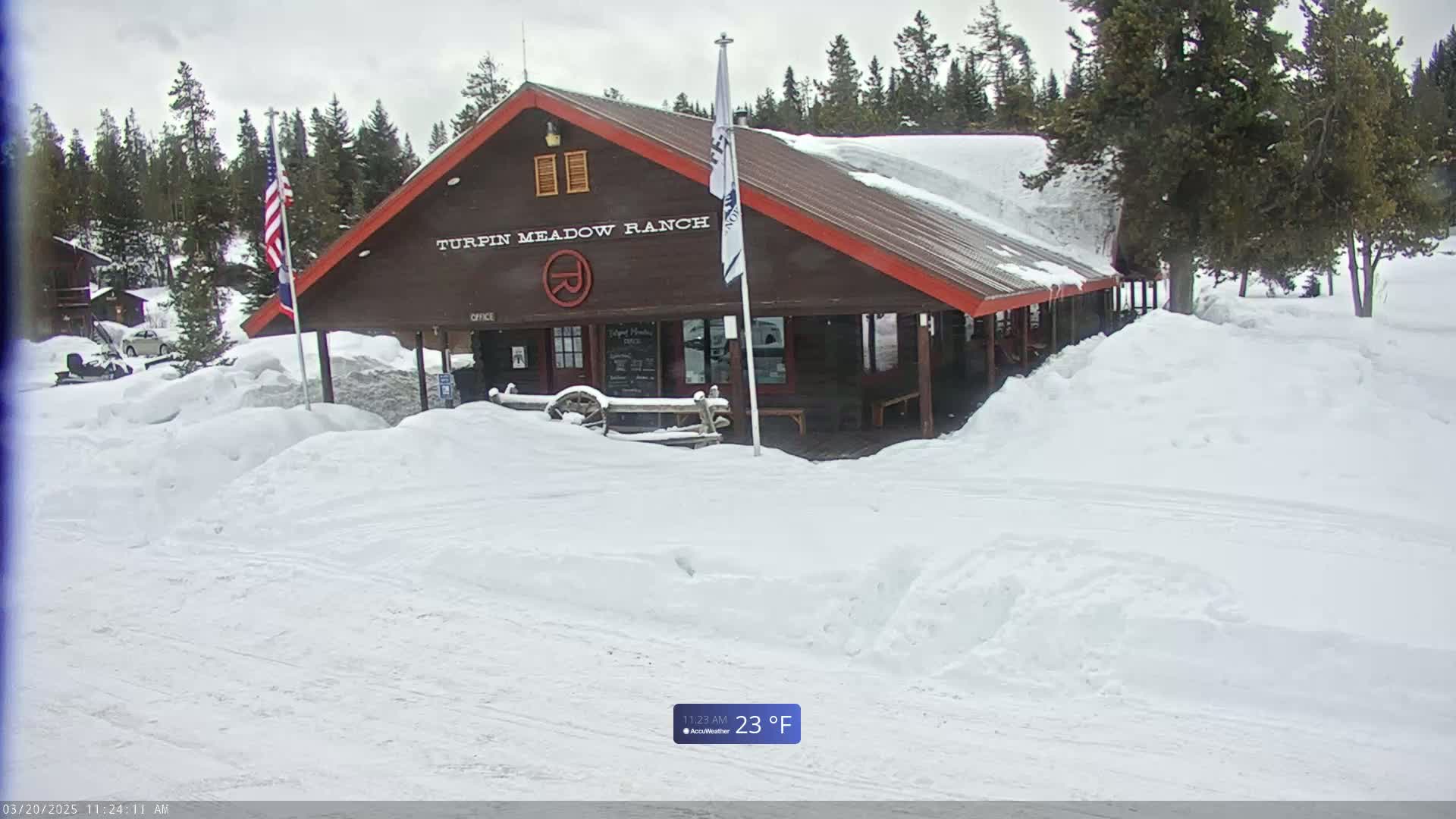 A snow-covered brown building with a red roof, surrounded by deep snow drifts, sits in a snowy landscape with evergreen trees under a cloudy sky.