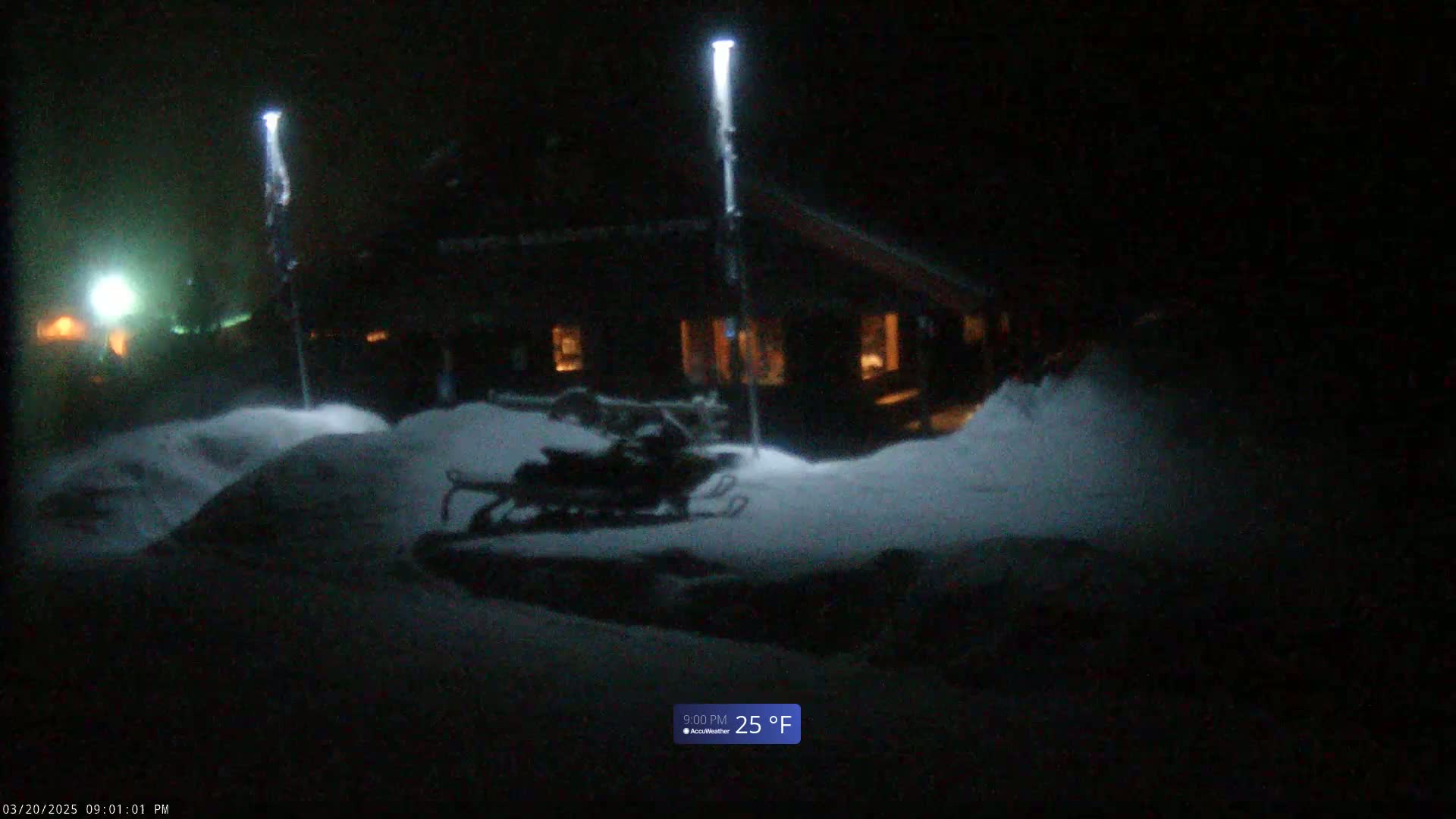 A snow-covered scene at night shows a dark building illuminated by tall lights, with a snowmobile parked nearby, under a dark, possibly foggy sky.