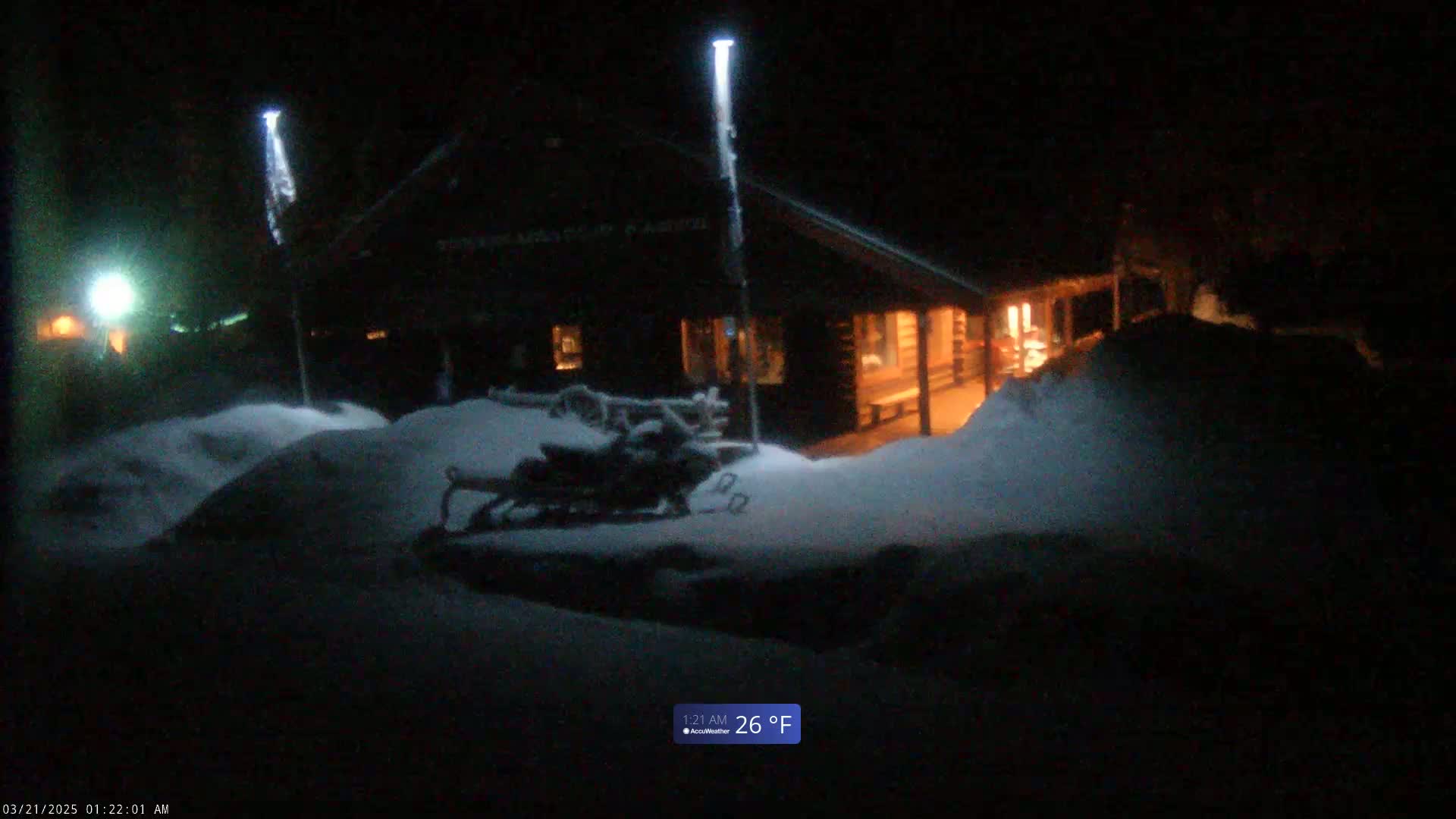 A snow-covered snowmobile sits outside a lit-up log cabin at night in the snow.