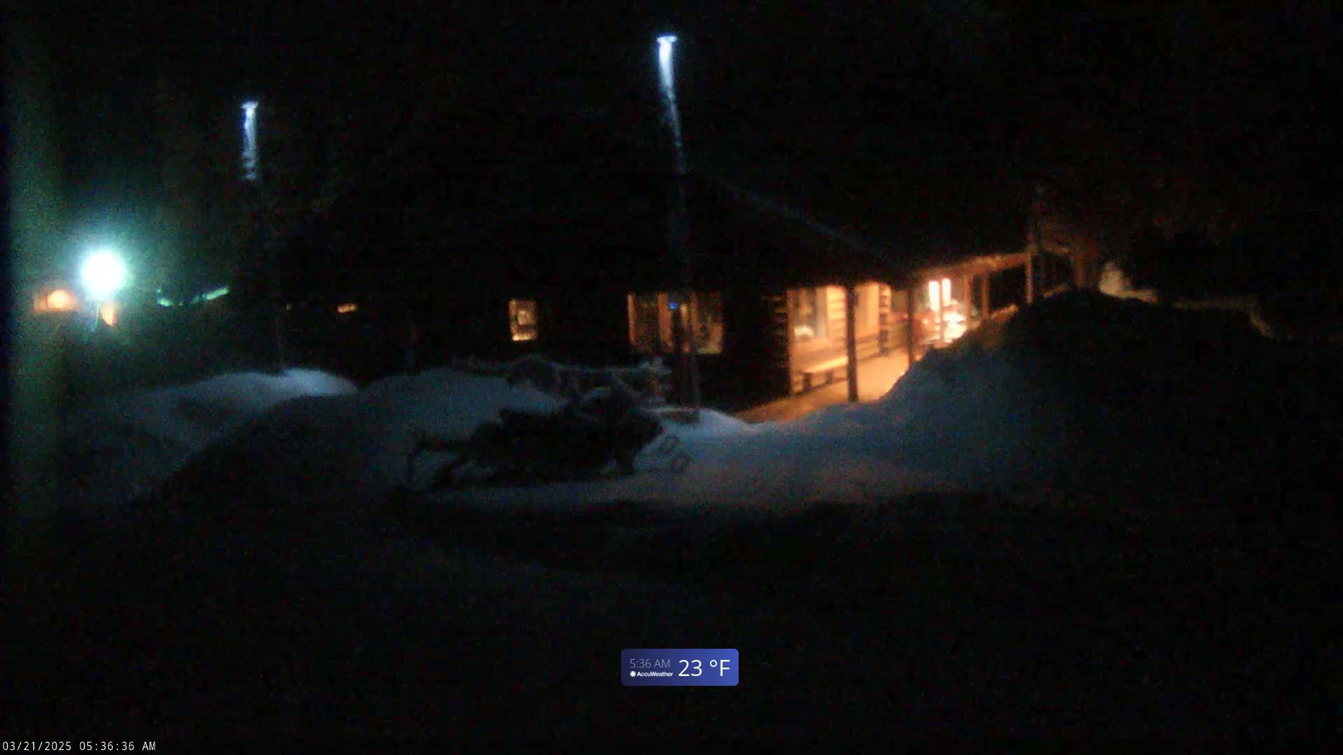 A snow-covered scene at night shows a dark log cabin illuminated from within, with a snowmobile partially visible in the foreground under bright lights.
