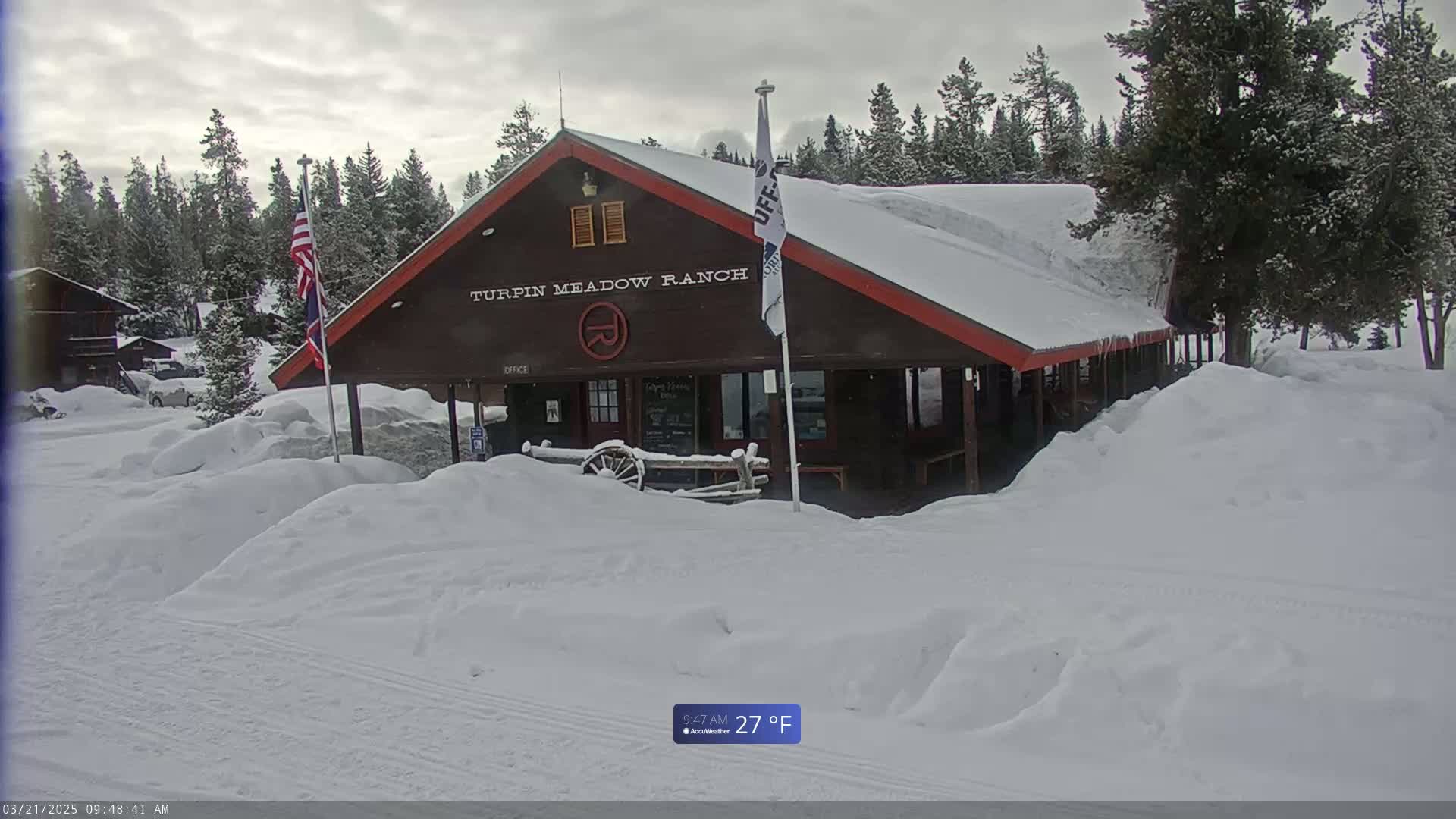 A snow-covered brown building with red trim,  surrounded by deep snow, under a cloudy sky.