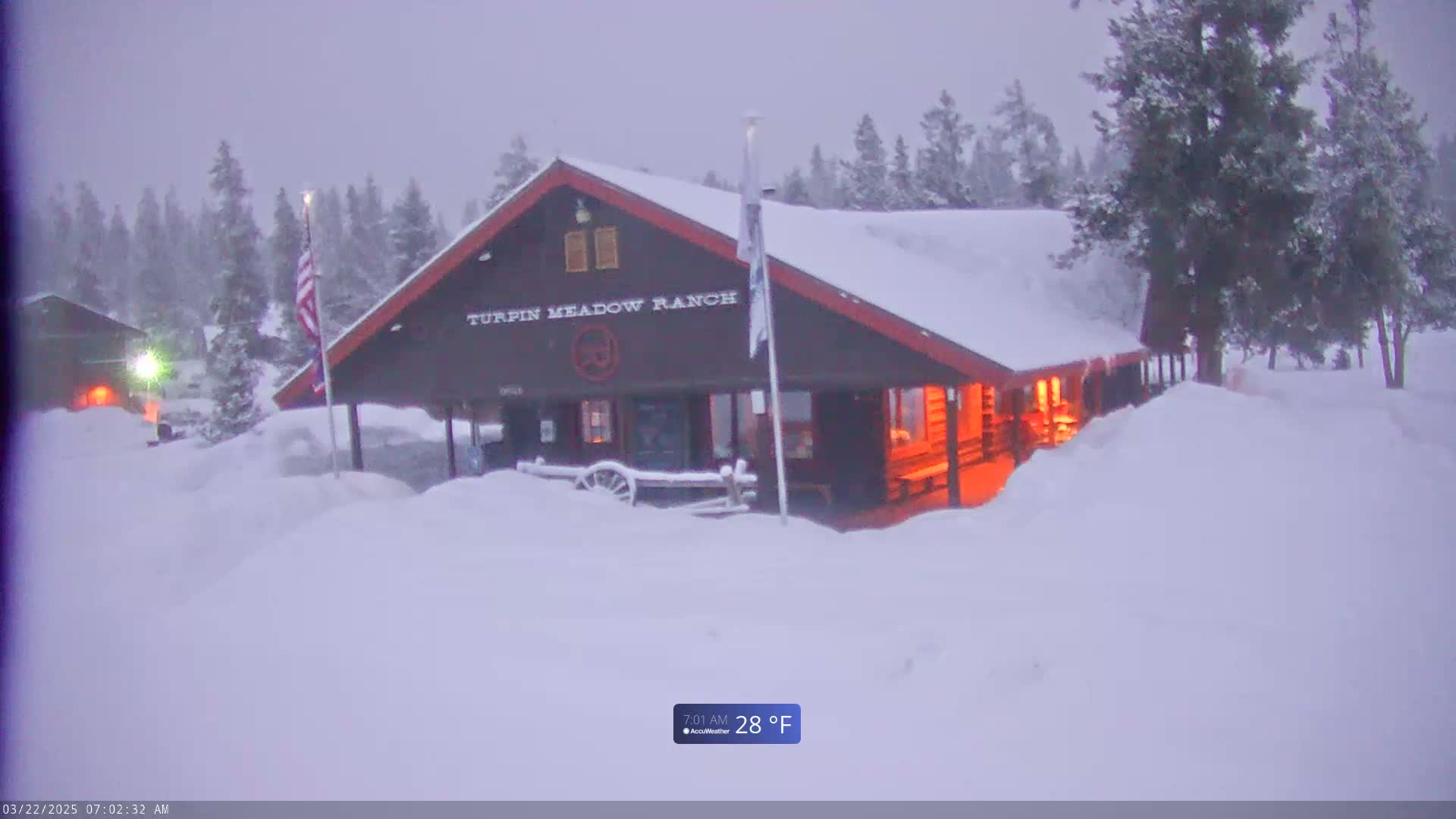 A snow-covered Turpin Meadow Ranch building is illuminated from within, surrounded by snow-covered trees in a snowy, overcast landscape.