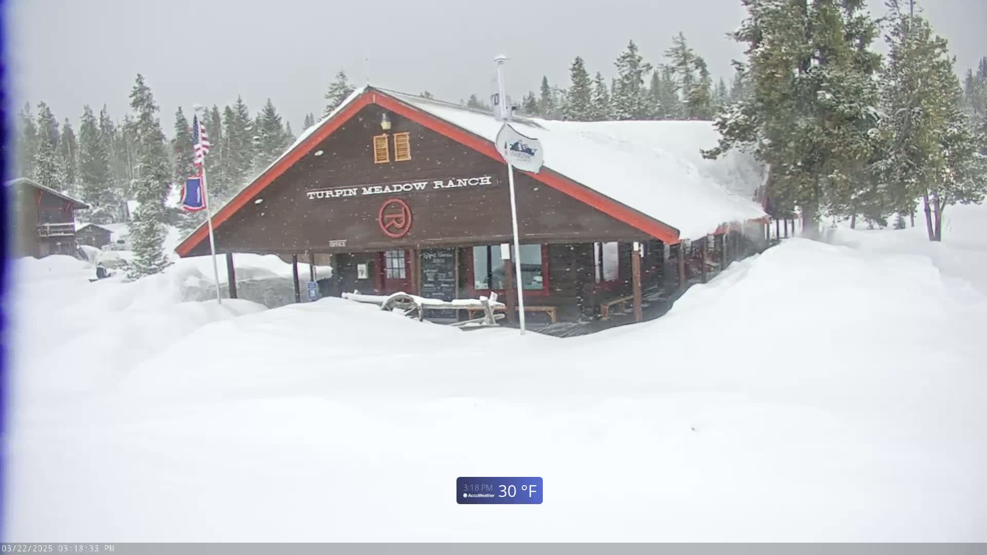 A brown building with a red roof is covered in snow during a snowfall, surrounded by deep snow drifts and a snowy forest in the background.