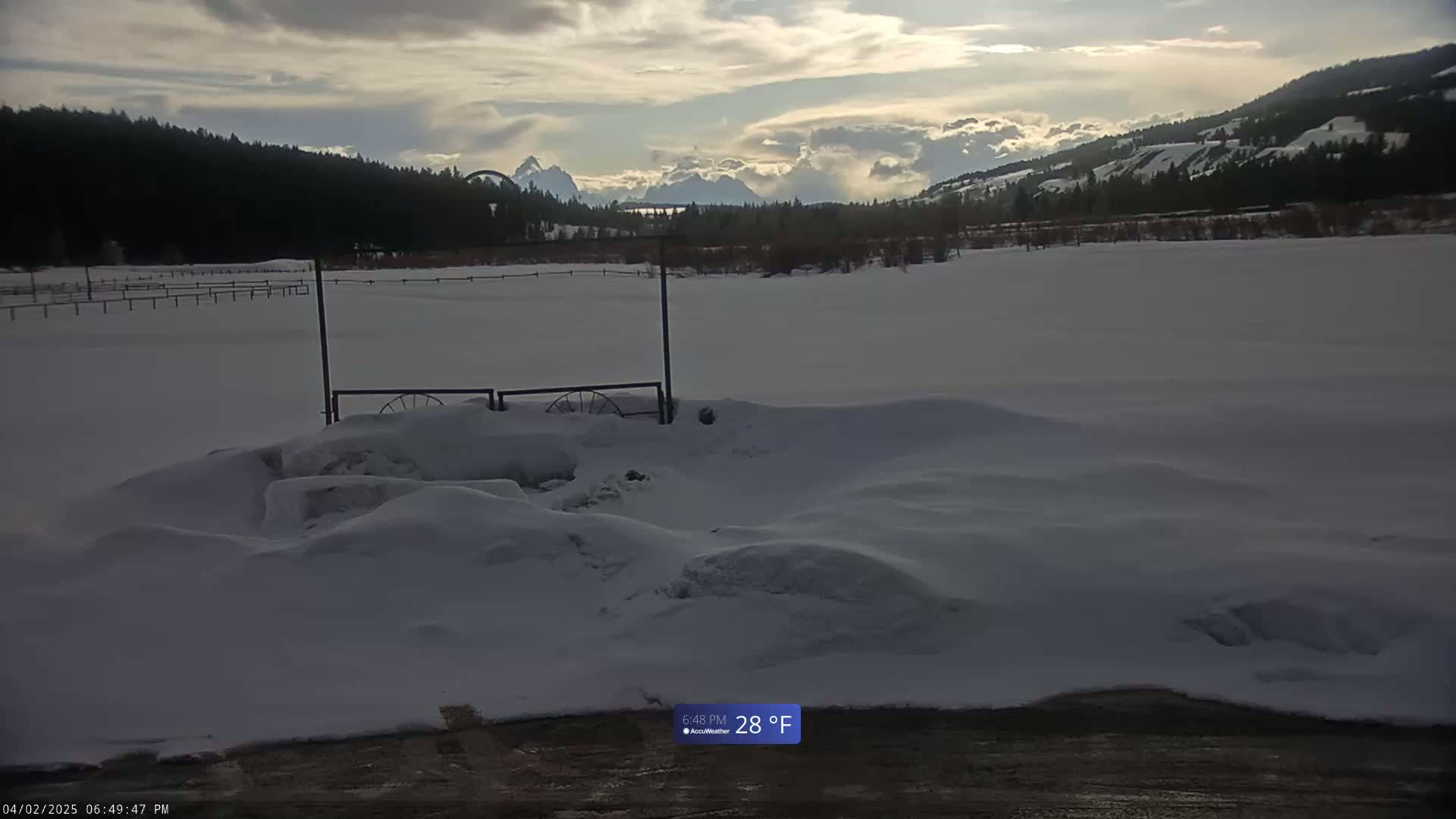 A snow-covered field with a gate and distant mountains under a cloudy sky.