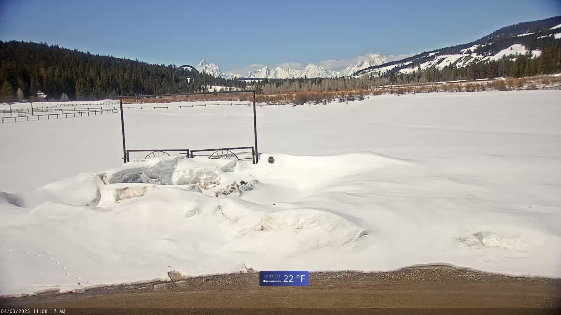 A snow-covered field with a partially buried gate and distant snow-capped mountains under a clear blue sky.