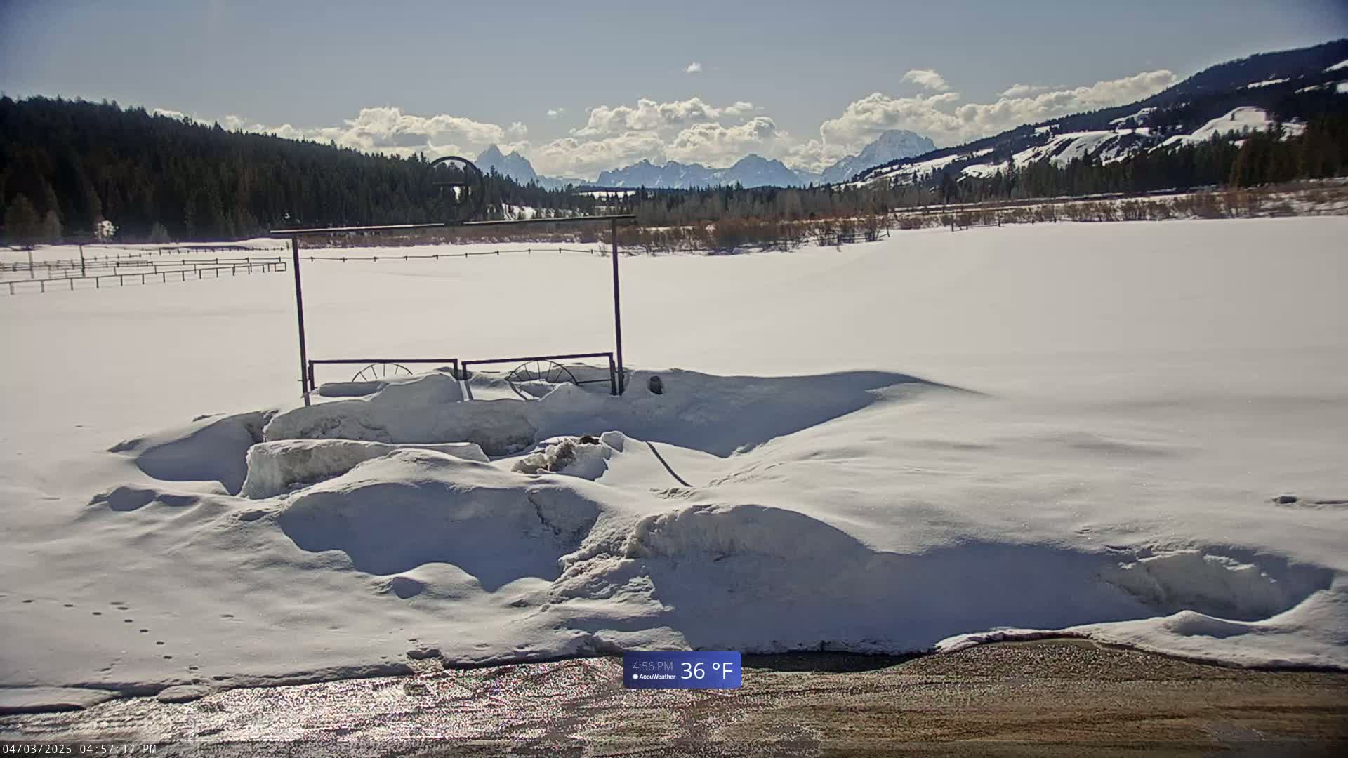 A snow-covered field with a metal gate and distant mountains under a partly cloudy sky.