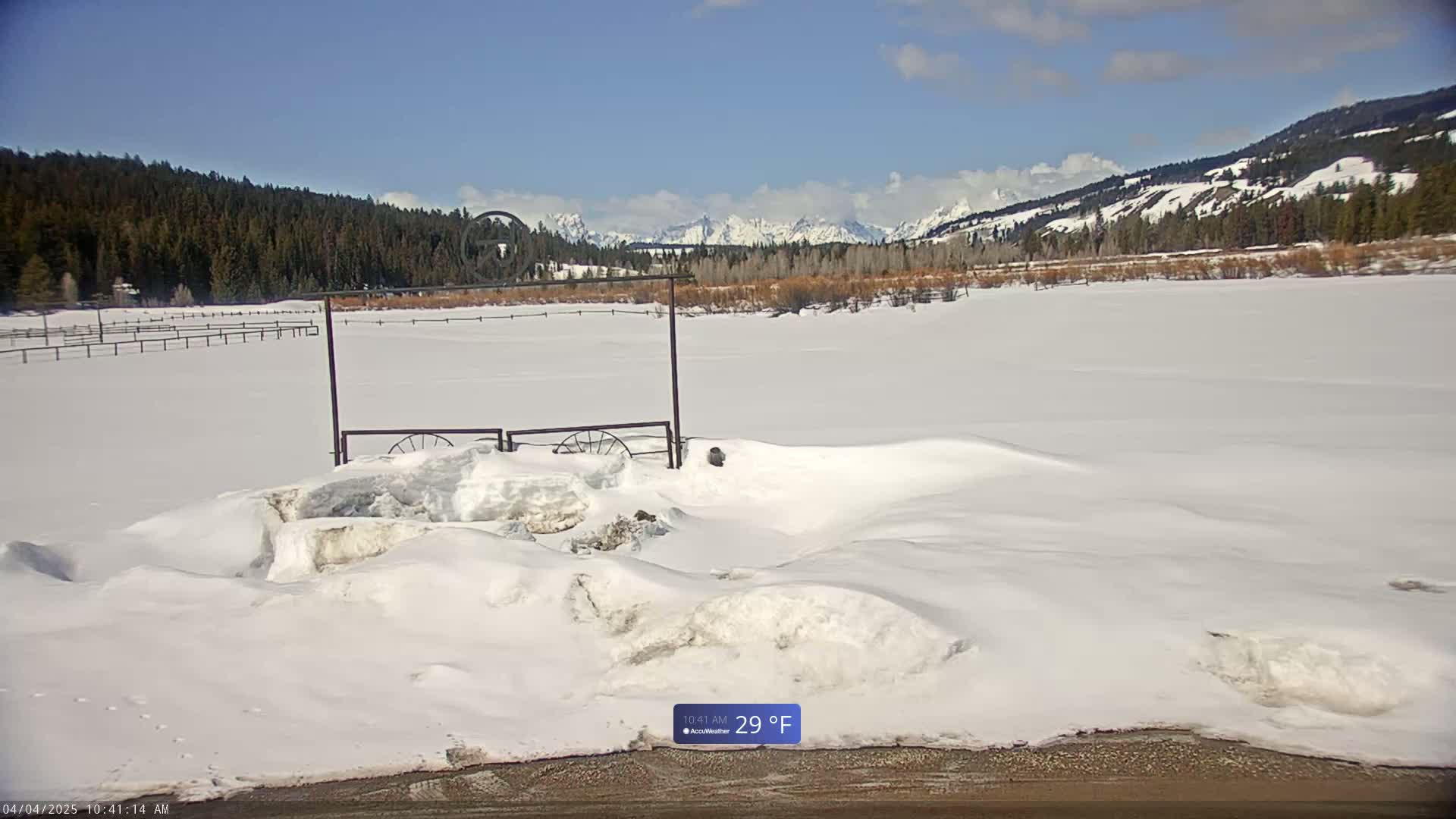 A snow-covered field with a metal gate and distant mountains under a mostly sunny sky.