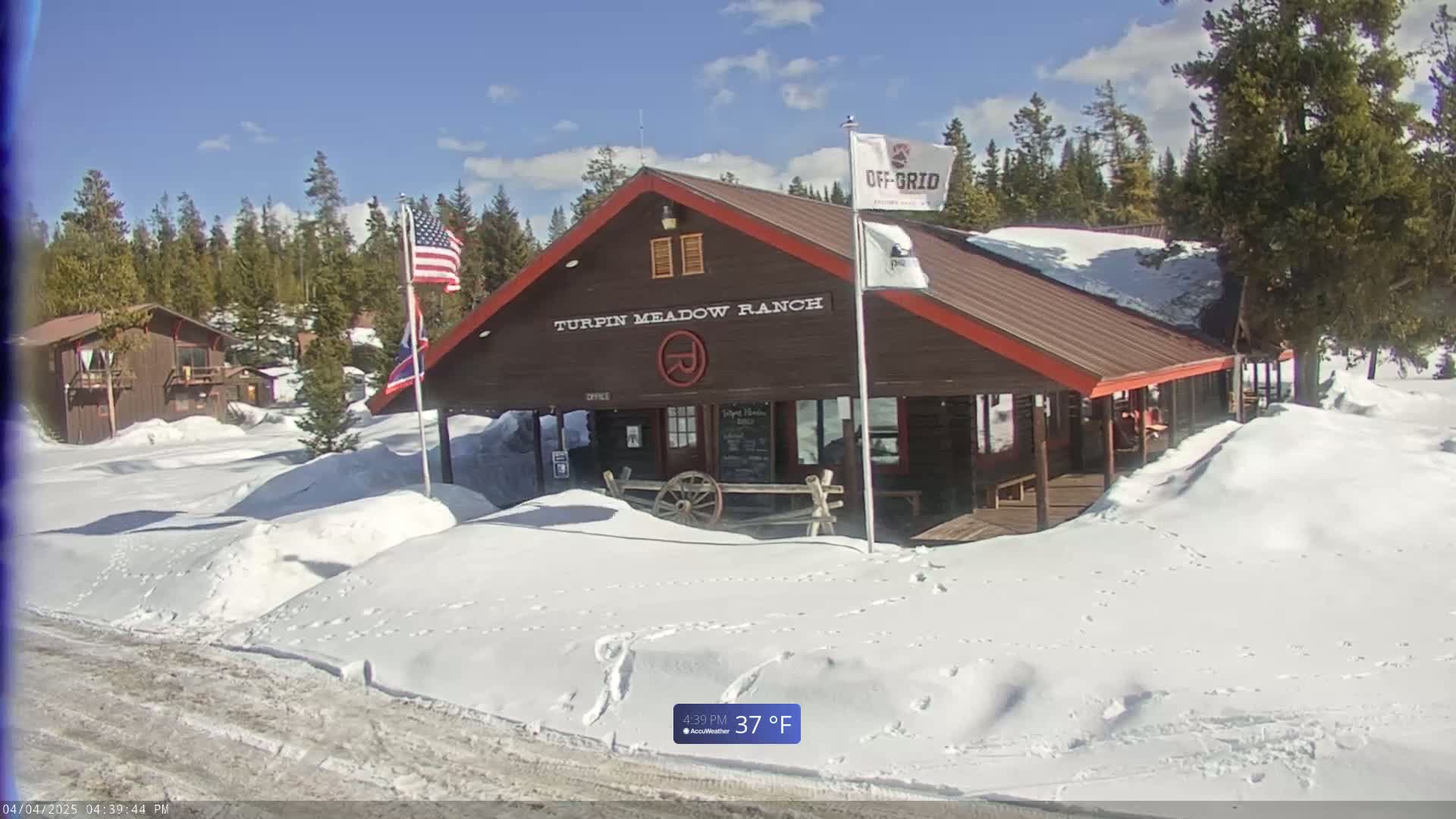 A snow-covered ranch building with a wooden porch and American and another flag is seen under a sunny sky.