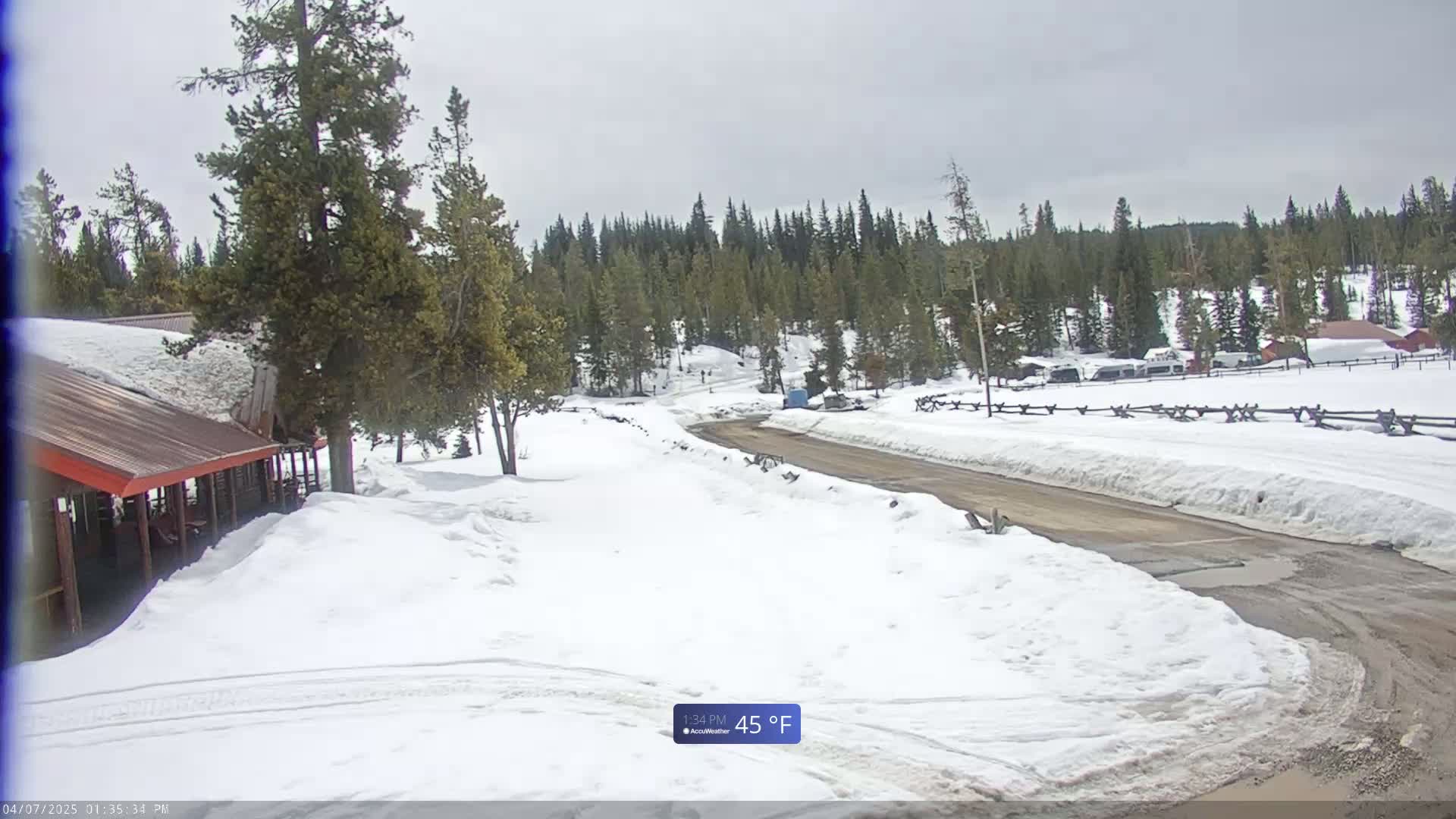 A snow-covered road winds through a pine forest towards a cluster of buildings under a cloudy sky.