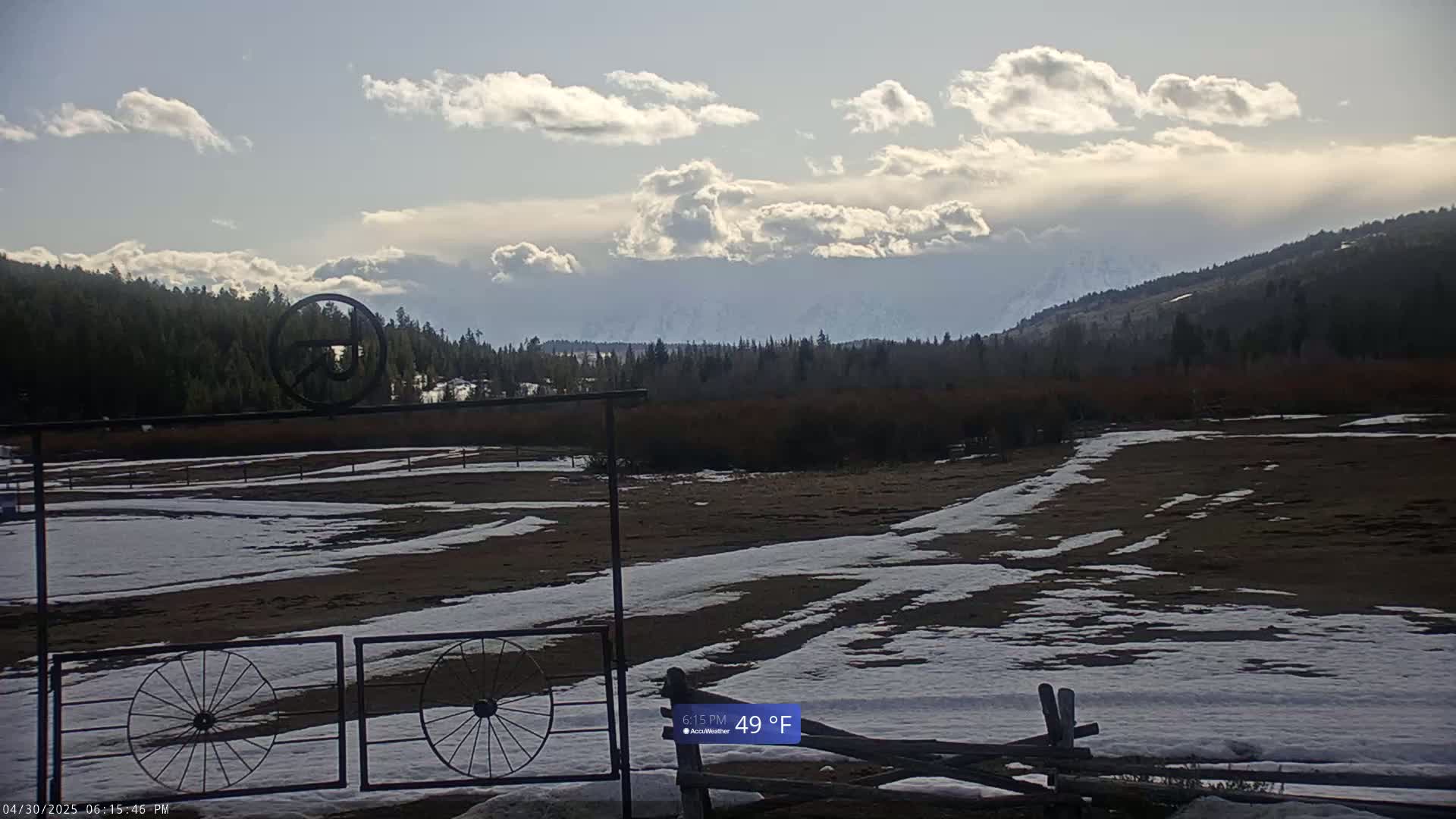 A partly cloudy sky hangs over a snow-dusted field, bordered by dark evergreen trees and distant mountains.
