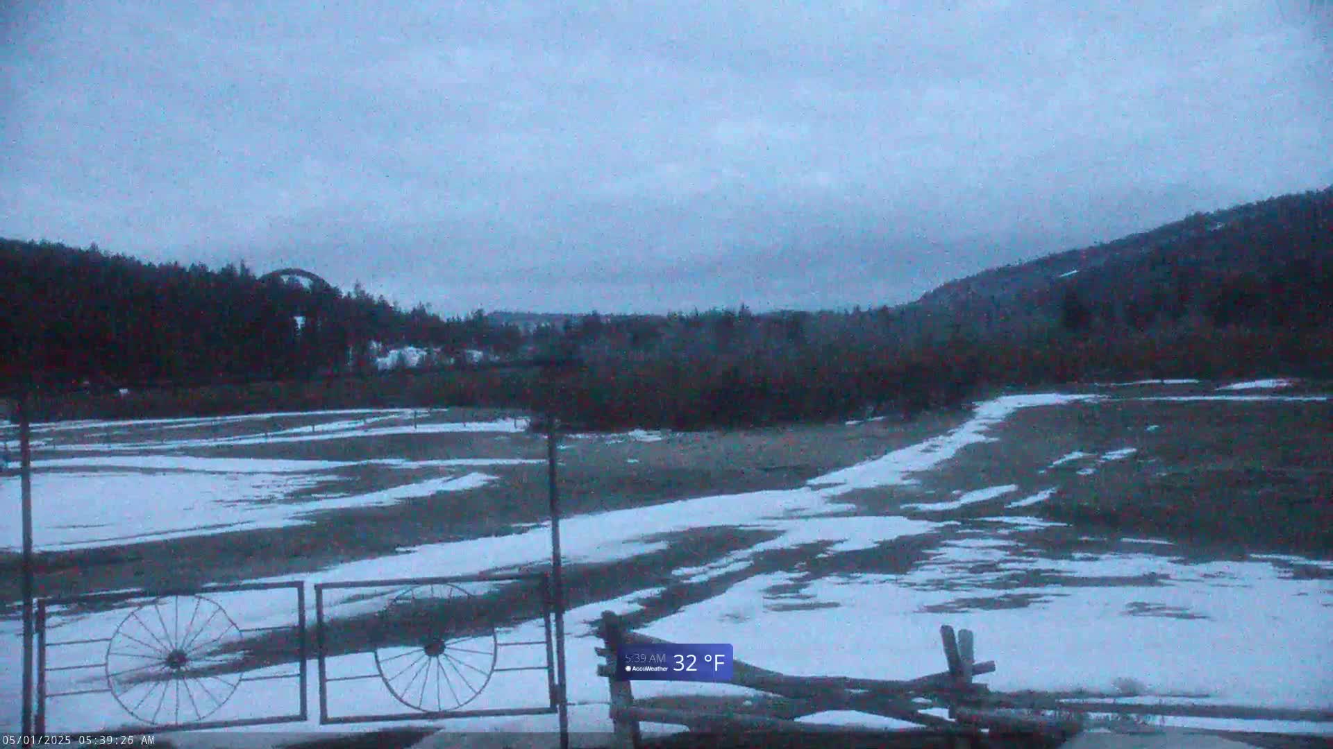 A snow-dusted field, partially covered in patchy snow, extends to a line of dark trees under a gray, overcast sky, with a temperature of 32°F.