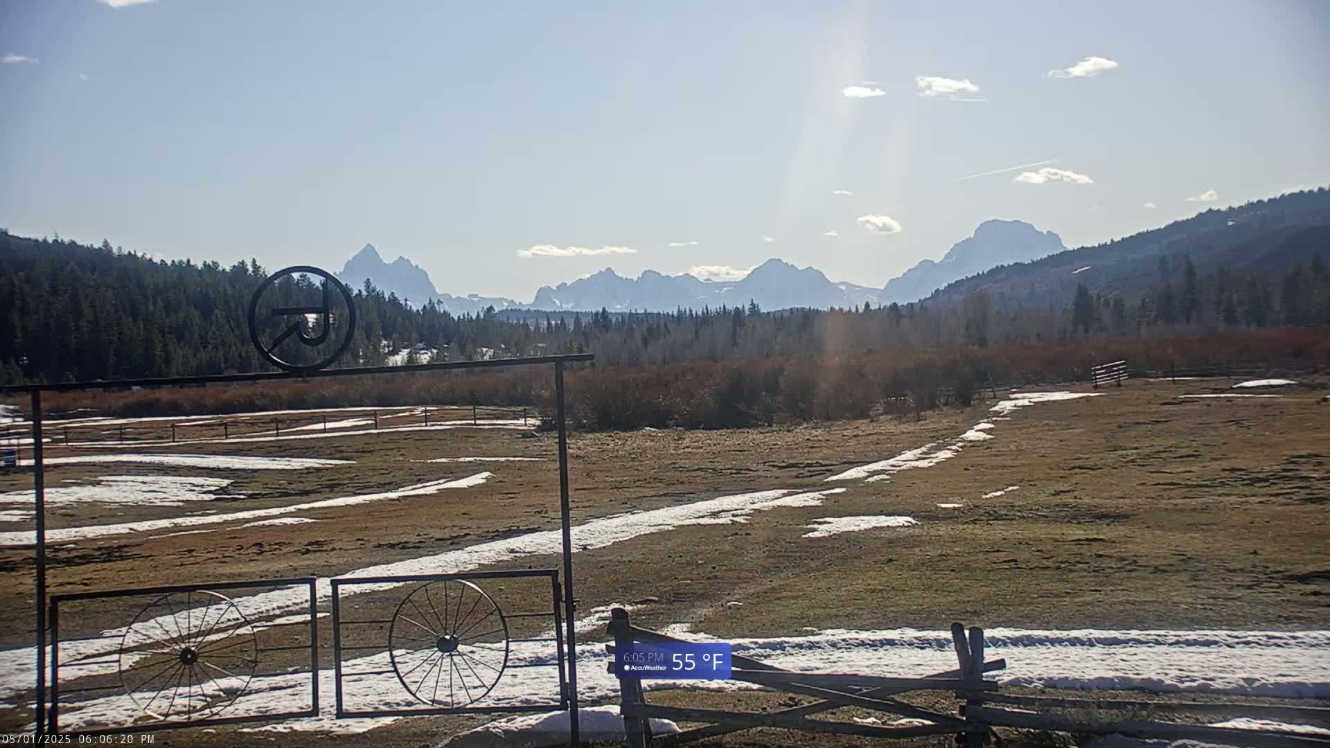 A partially snow-covered field with a metal gate featuring wagon wheels sits before a backdrop of a mountain range under a partly sunny sky.