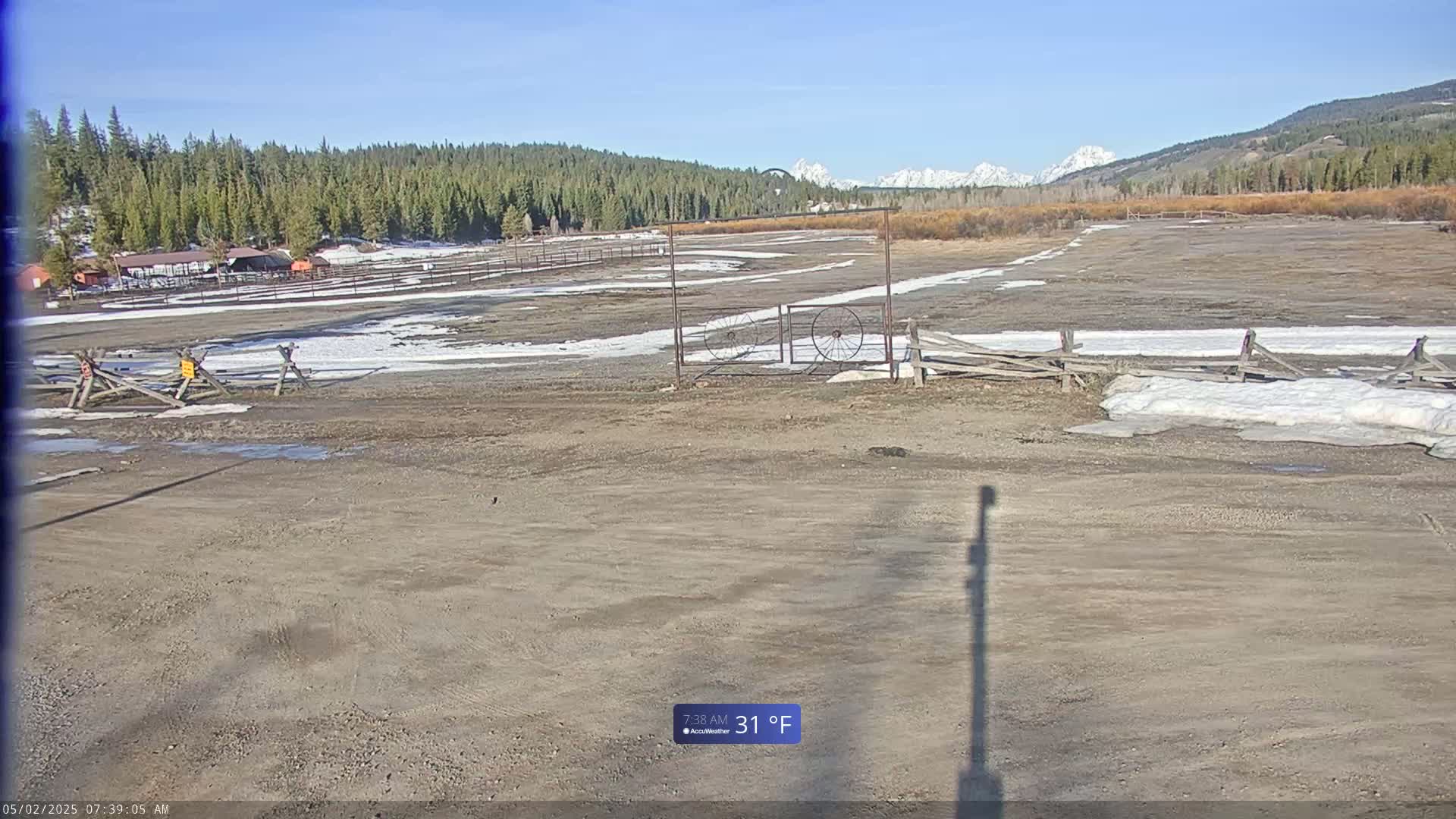 A mostly dirt field with patches of snow and a wooden fence, sits in front of a forest with snow-capped mountains in the background on a sunny, 31°F day.