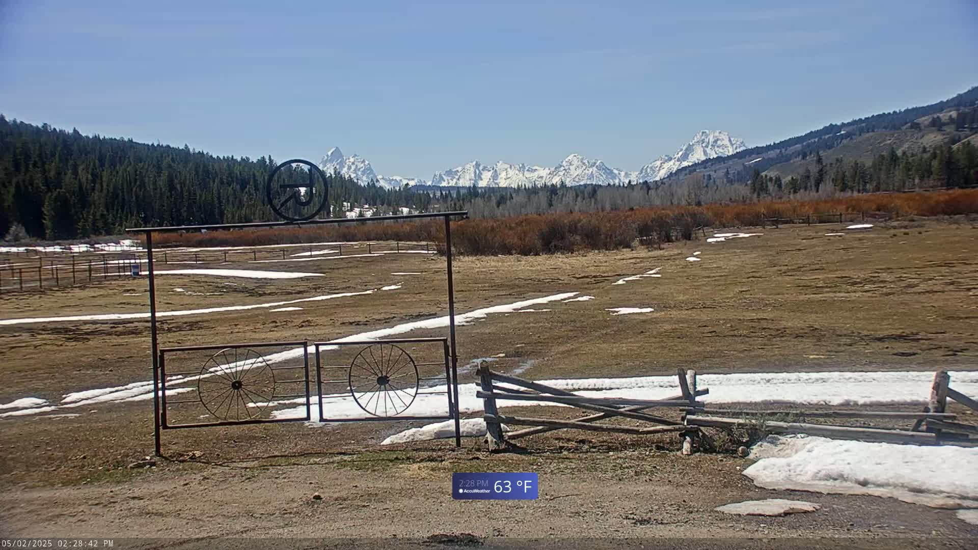 A snow-dusted field with a rustic metal gate and wooden fence stretches towards a mountain range under a clear blue sky.