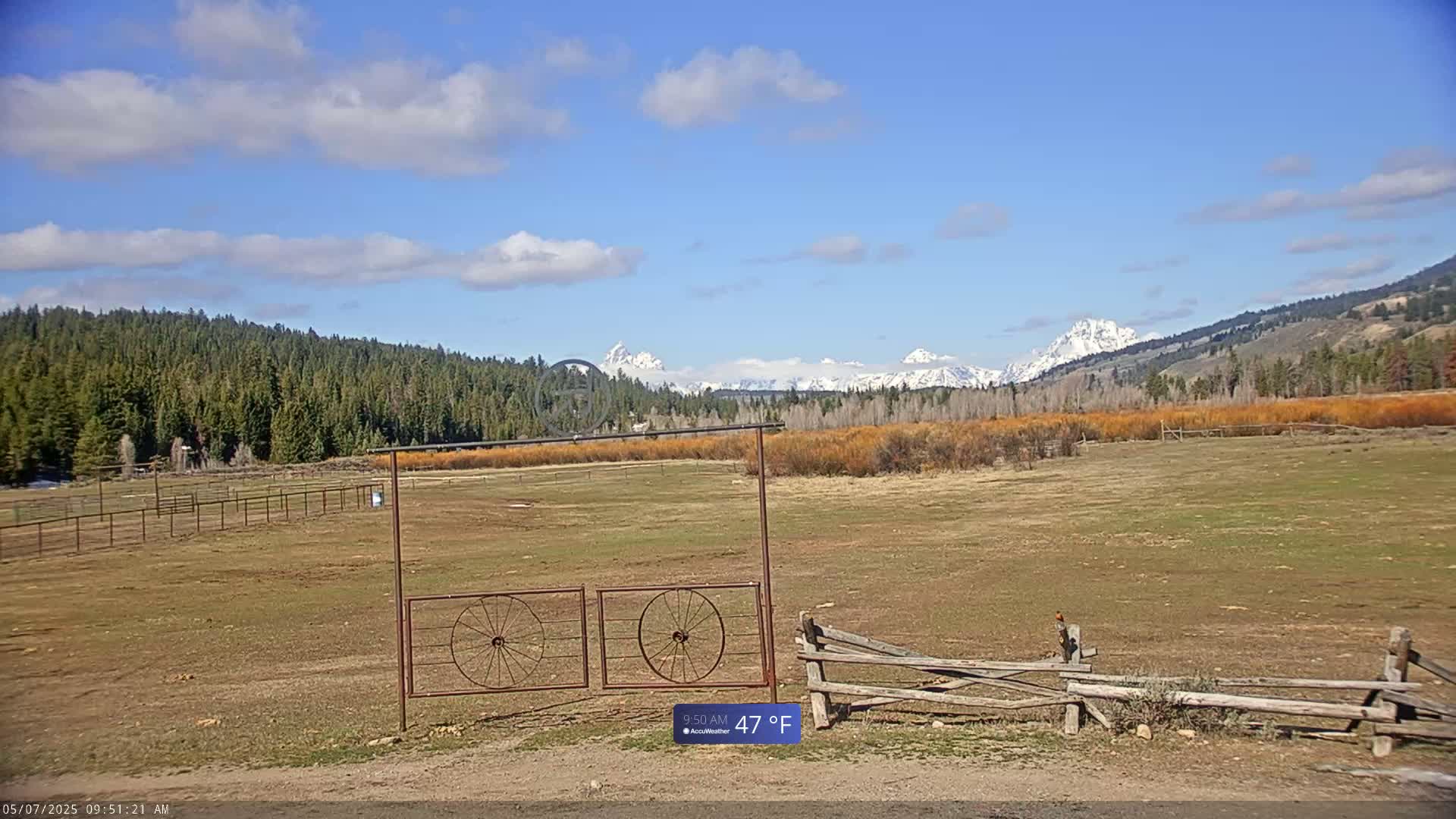 A partly cloudy, 47°F day reveals a snow-capped mountain range in the distance, behind a field with a wooden fence and a metal gate featuring wagon wheel designs.