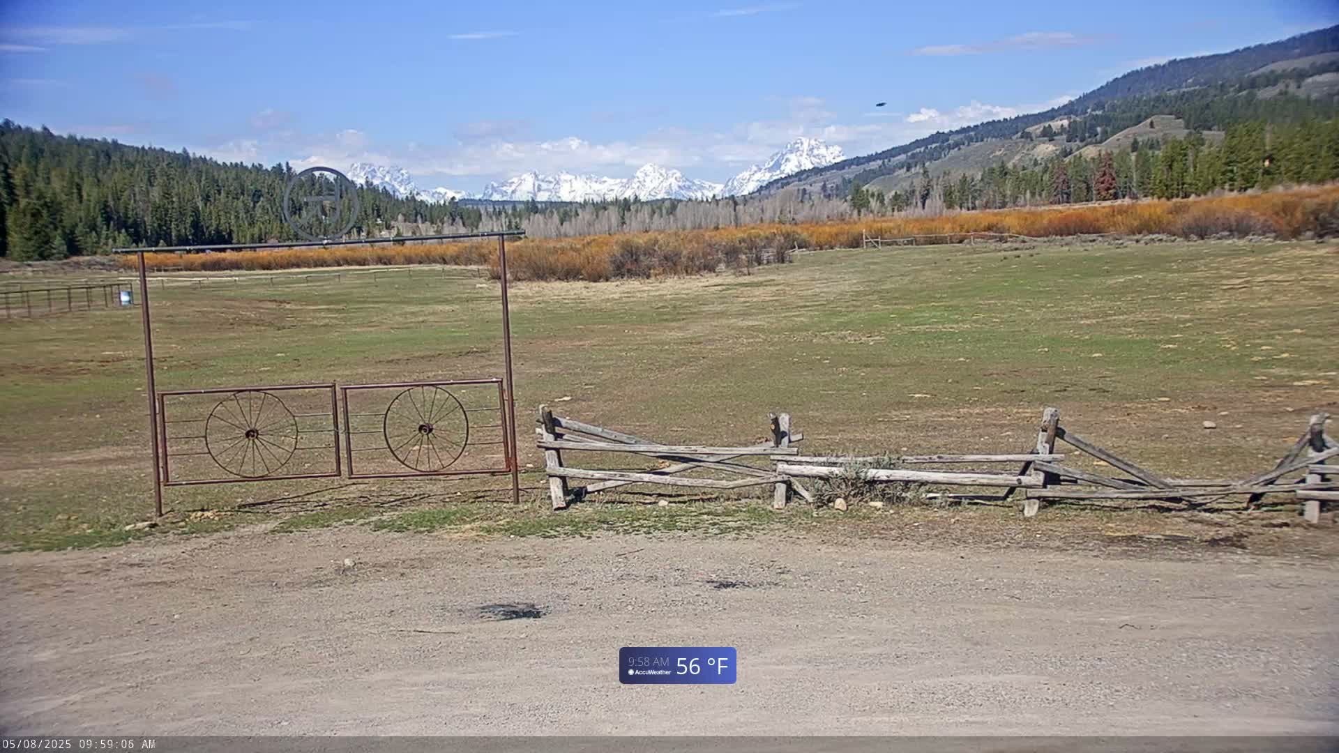 A grassy field with a split-rail fence and metal gate in the foreground, snow-capped mountains in the background under a clear blue sky.