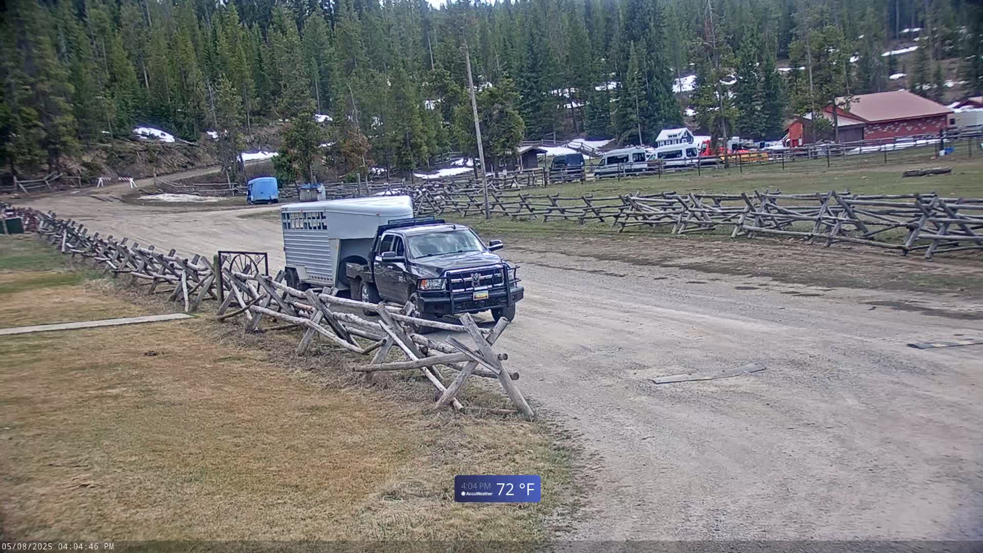 A dark-colored pickup truck towing a horse trailer travels on a dirt road past split-rail fences, with a background of pine trees and buildings, on a partly sunny 72°F day.