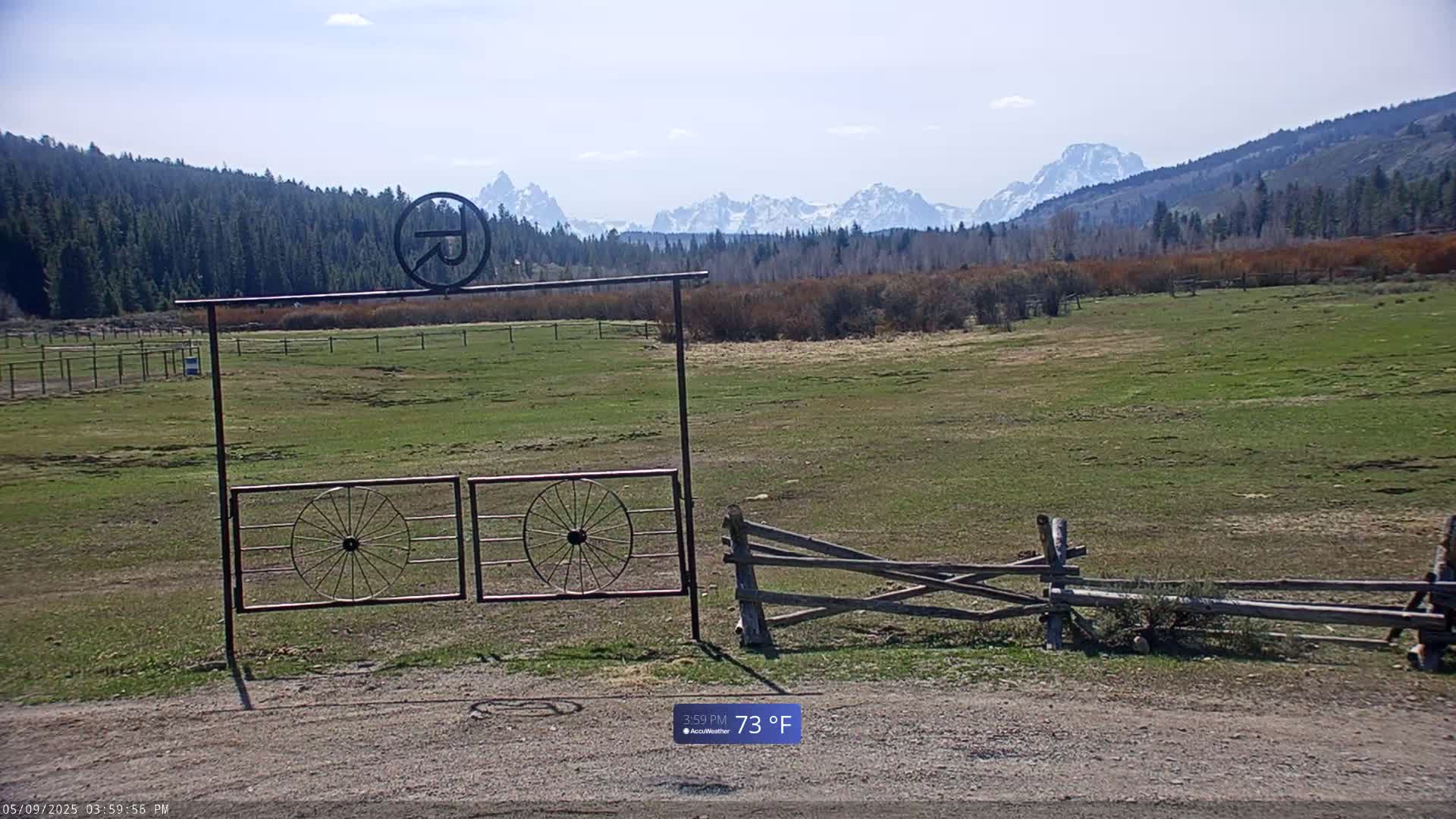 A metal gate with wagon wheel designs sits in front of a grassy field, beyond which are snow-capped mountains under a mostly clear sky.