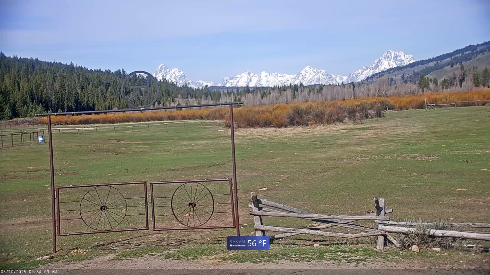 A rusty metal gate with wagon wheel designs sits in front of a grassy field, with a snow-capped mountain range in the background under a clear blue sky.