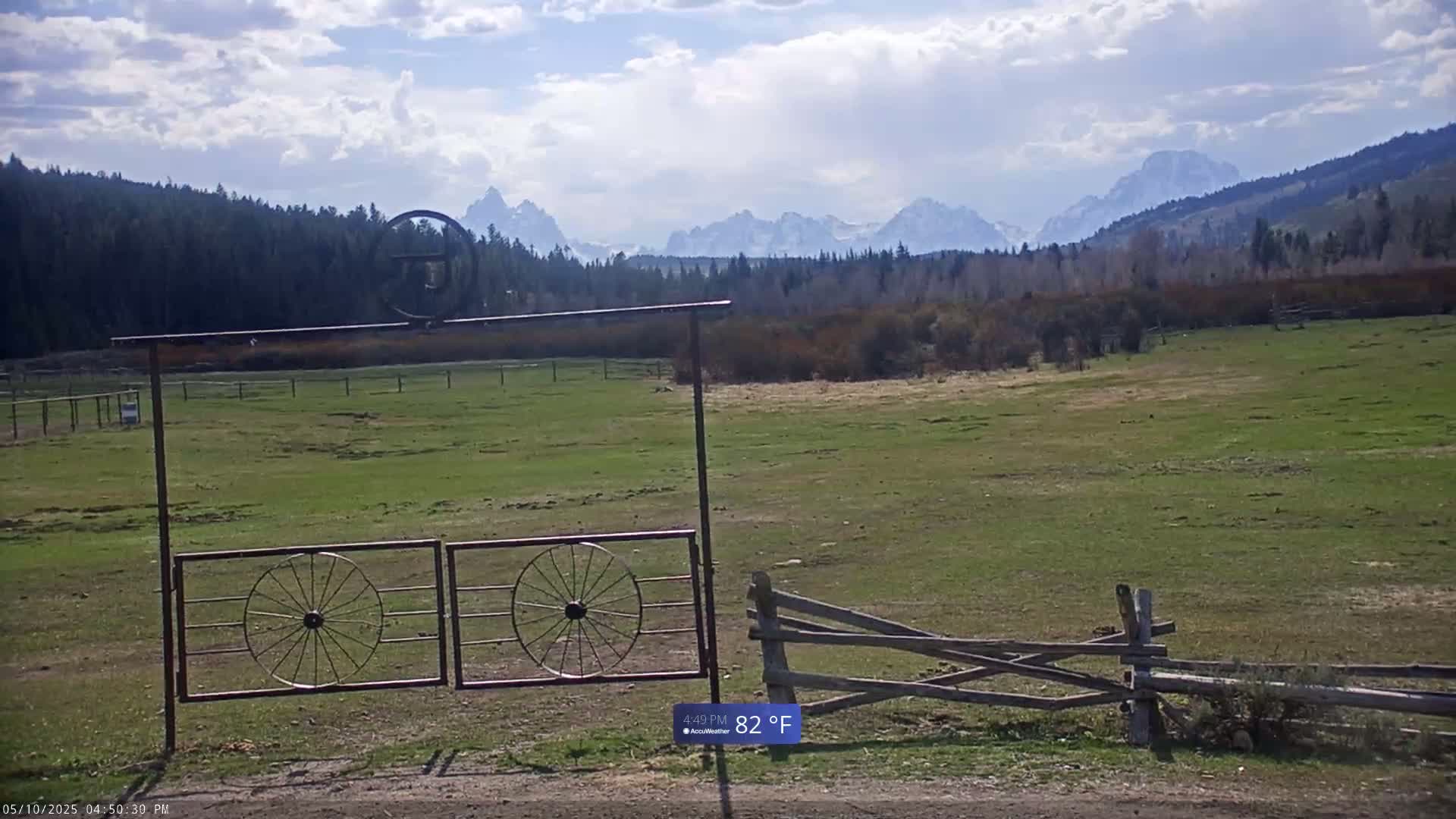 A partially cloudy sky overlooks a grassy field with a metal gate featuring wagon wheels, a split-rail fence, and snow-capped mountains in the distance.