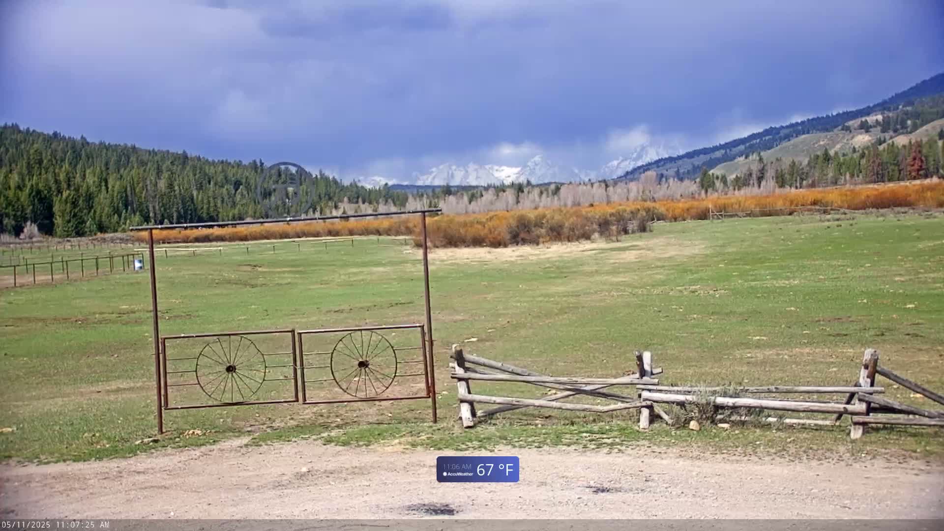 A grassy field with a rustic wooden fence and a metal gate, under a partly cloudy sky, overlooks a forested hillside and snow-capped mountains in the distance.