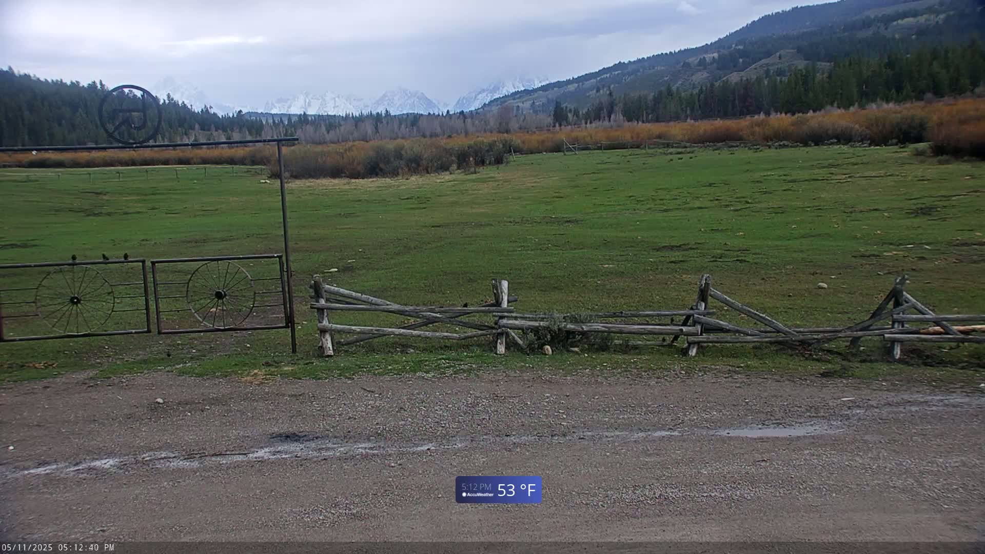 A grassy field with a split-rail fence and a metal gate featuring wagon wheels is visible, backed by a mountain range under a cloudy sky.