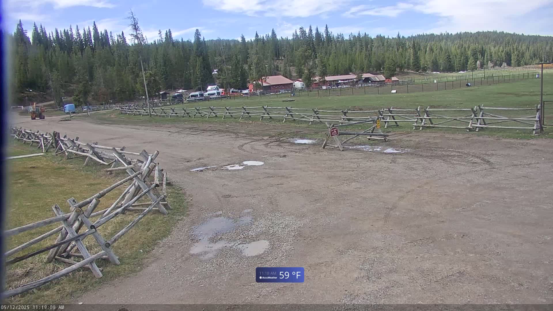 A dirt road with puddles leads to a grassy field enclosed by wooden split-rail fences, beyond which are several buildings nestled amongst a coniferous forest under a partly cloudy sky.