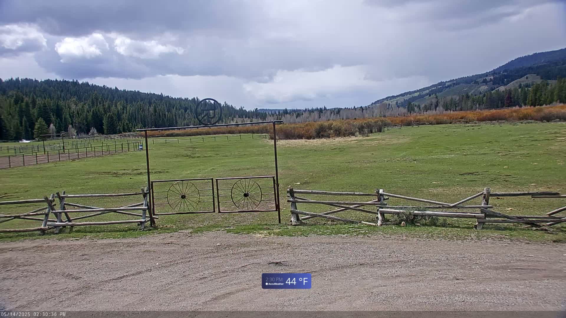 A grassy field bordered by a wooden split-rail fence and a metal gate with wagon wheel designs sits under a partly cloudy sky.