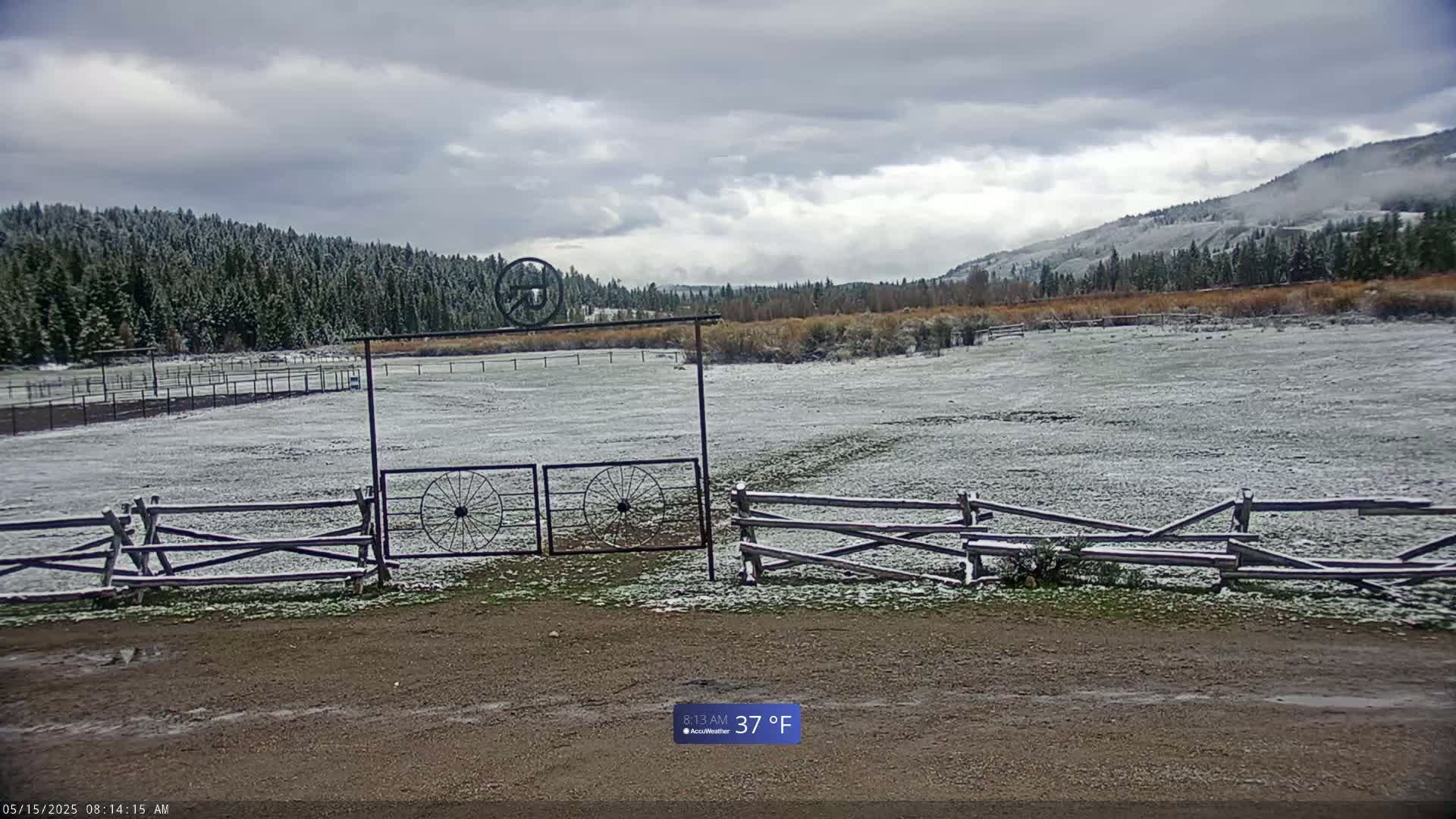 A snow-dusted field is enclosed by a wooden split-rail fence and an ornate metal gate, with a backdrop of a snow-covered evergreen forest and distant hills under a cloudy sky.