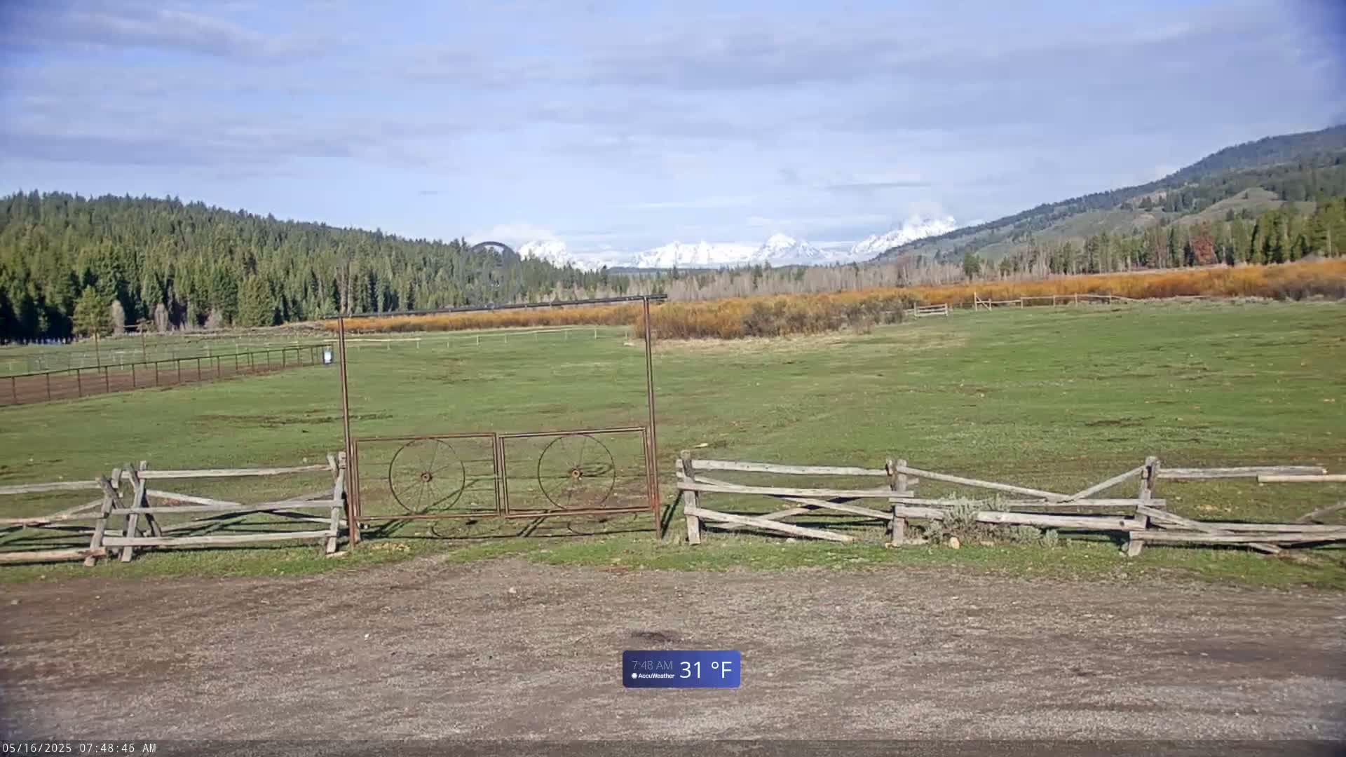 A grassy field with a split-rail fence and metal gate in the foreground, backed by a forest and snow-capped mountains under a partly cloudy sky.