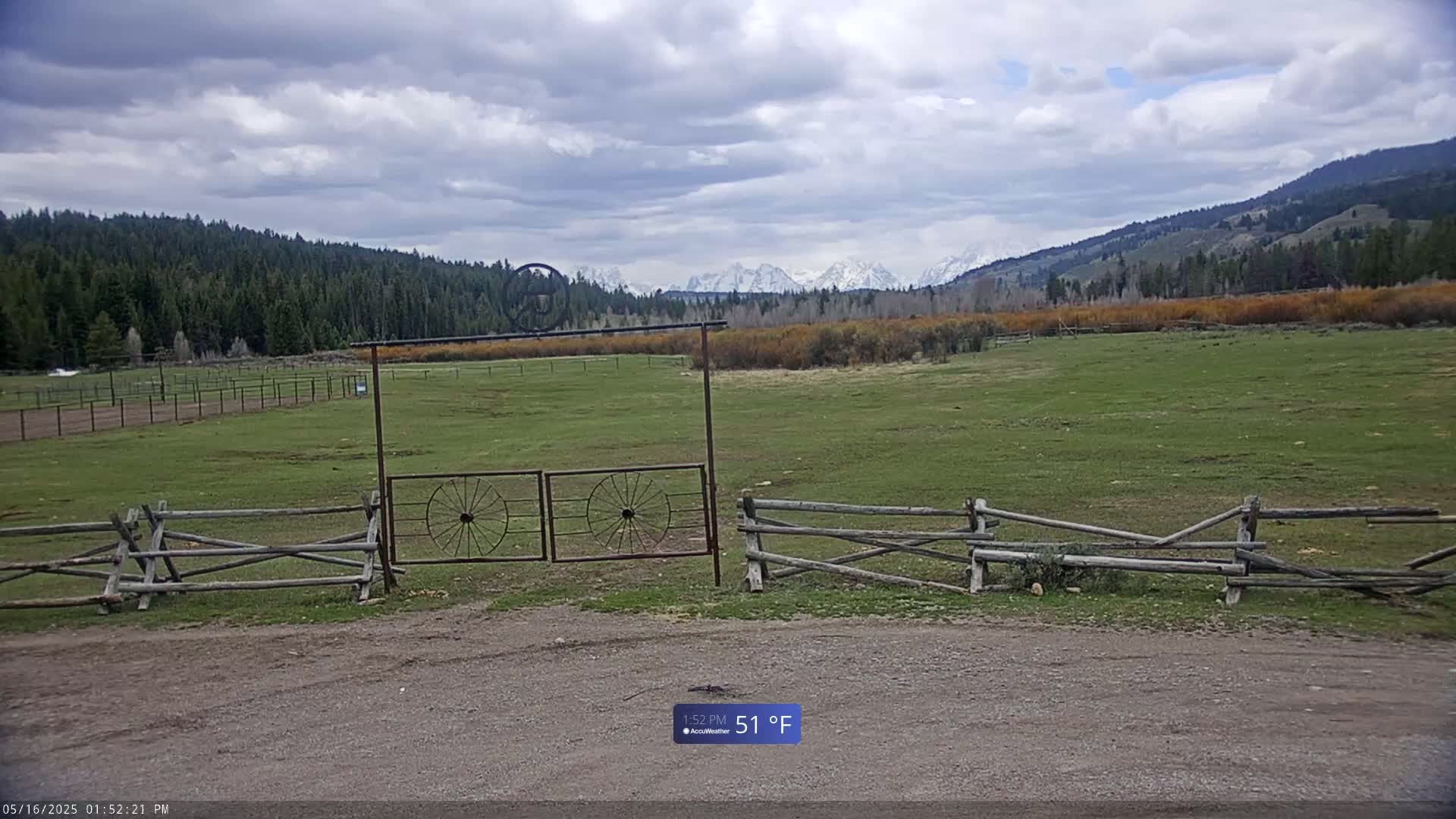 A grassy field with a split-rail fence and metal gate opens to a view of snow-capped mountains under a cloudy sky.