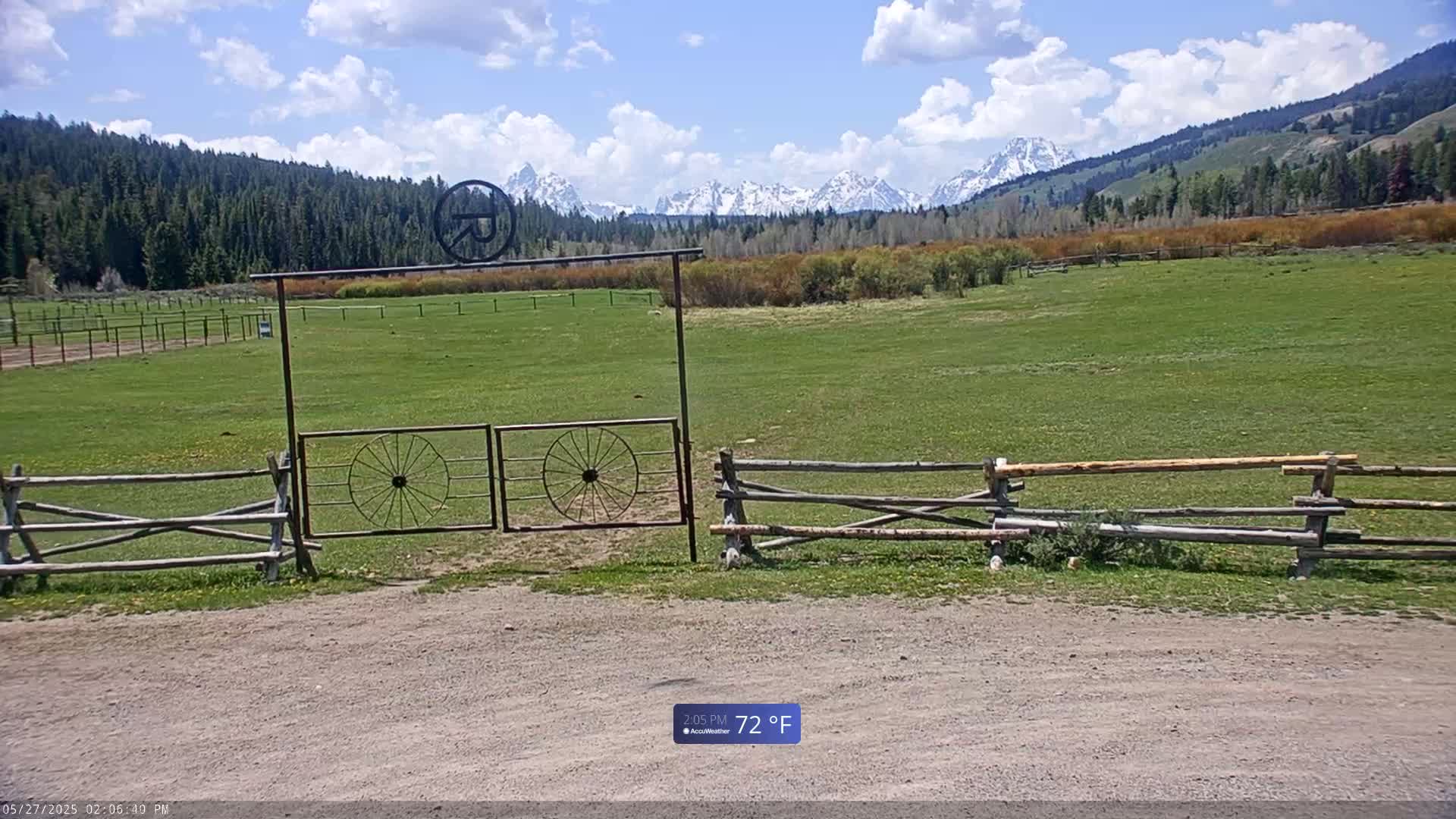 A grassy field with a split-rail fence and wagon wheel gate leads to snow-capped mountains under a partly cloudy sky.