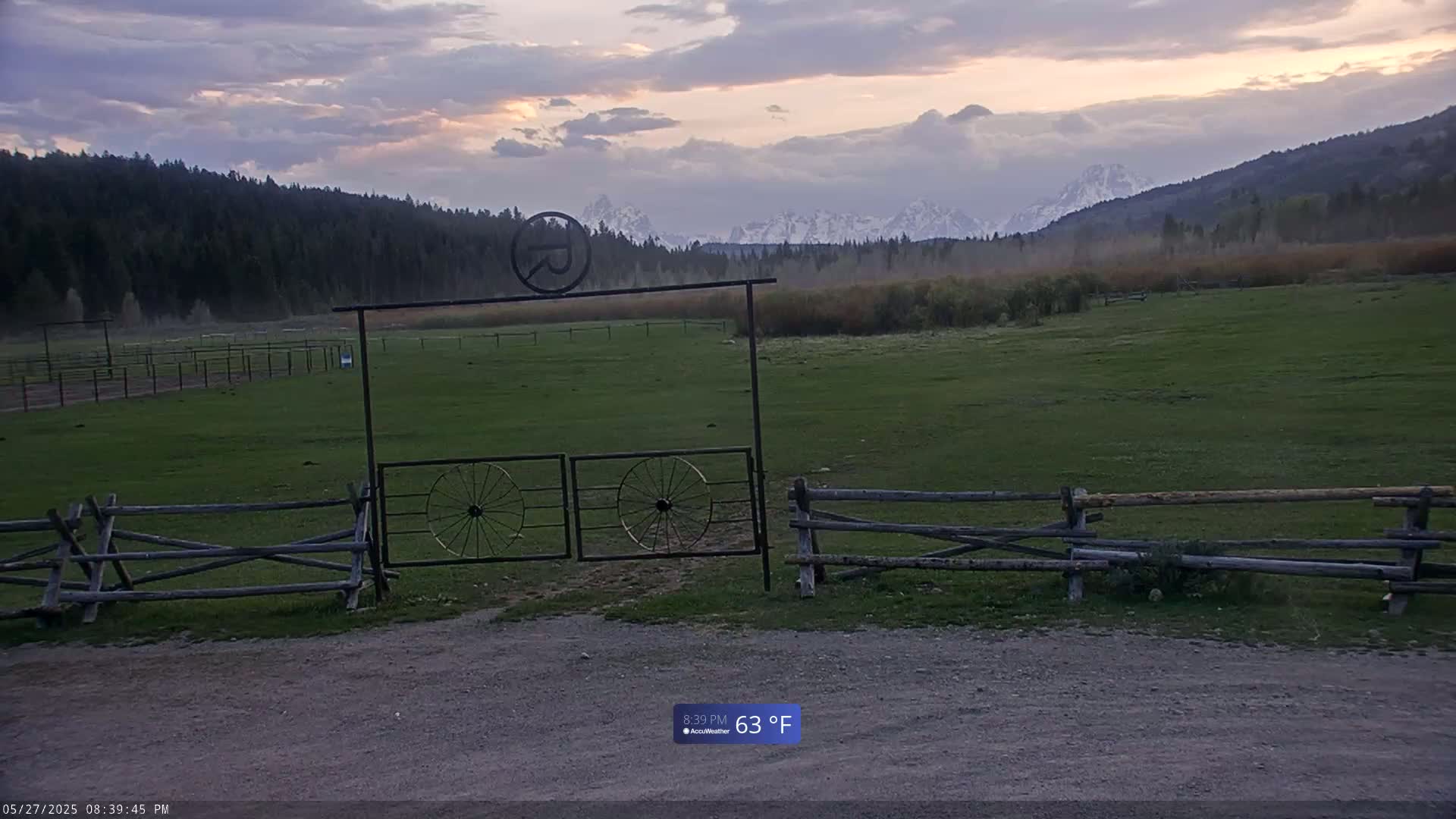 A grassy field with a wooden split-rail fence and a metal gate featuring wagon wheels, overlooks a hazy mountain range under a partly cloudy sky at sunset.