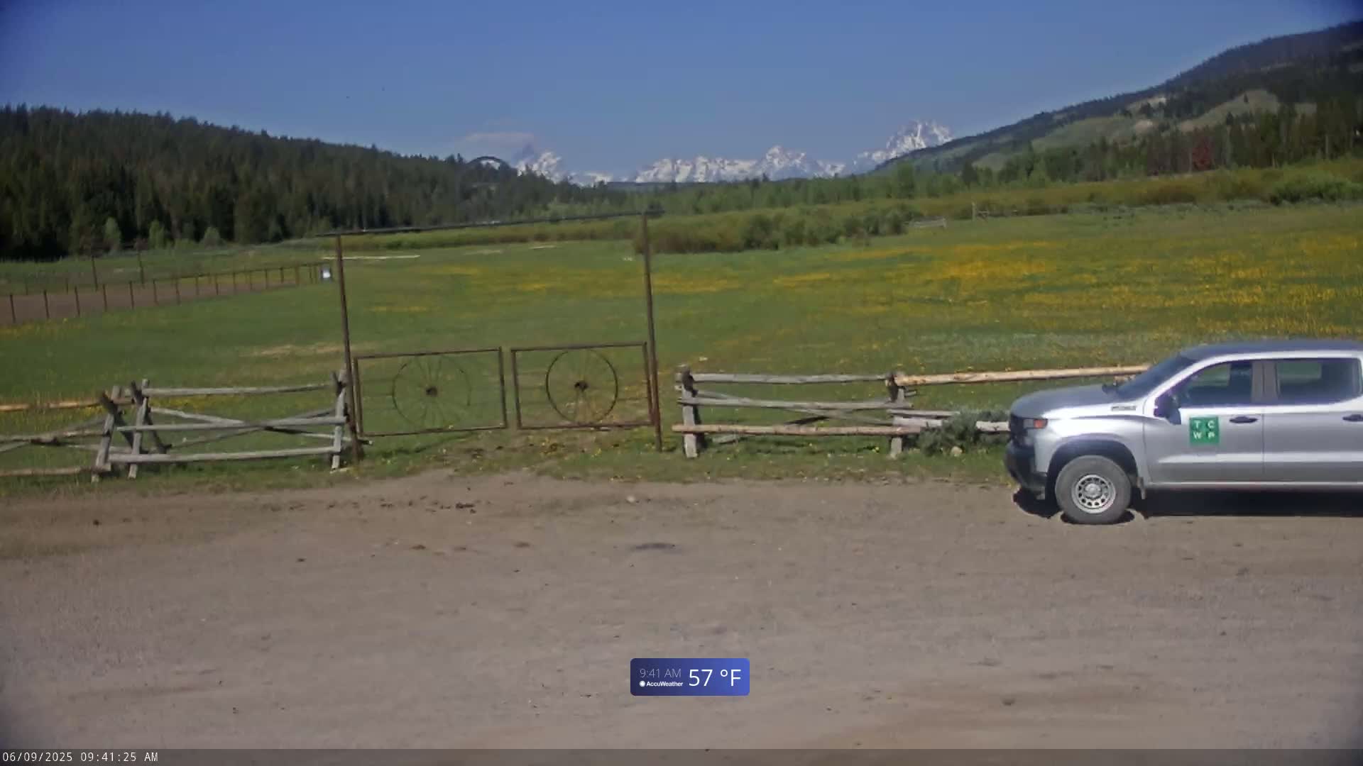 A silver pickup truck is parked near a wooden fence and gate in front of a field of yellow wildflowers, with mountains in the background on a sunny 57°F day.