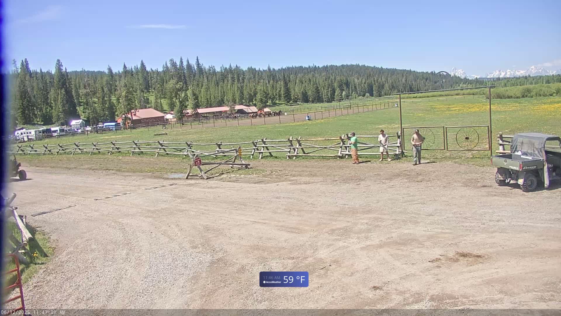 A dirt lot with three people standing near a wooden fence and gate, a utility vehicle parked nearby, and a ranch in the background on a sunny day.