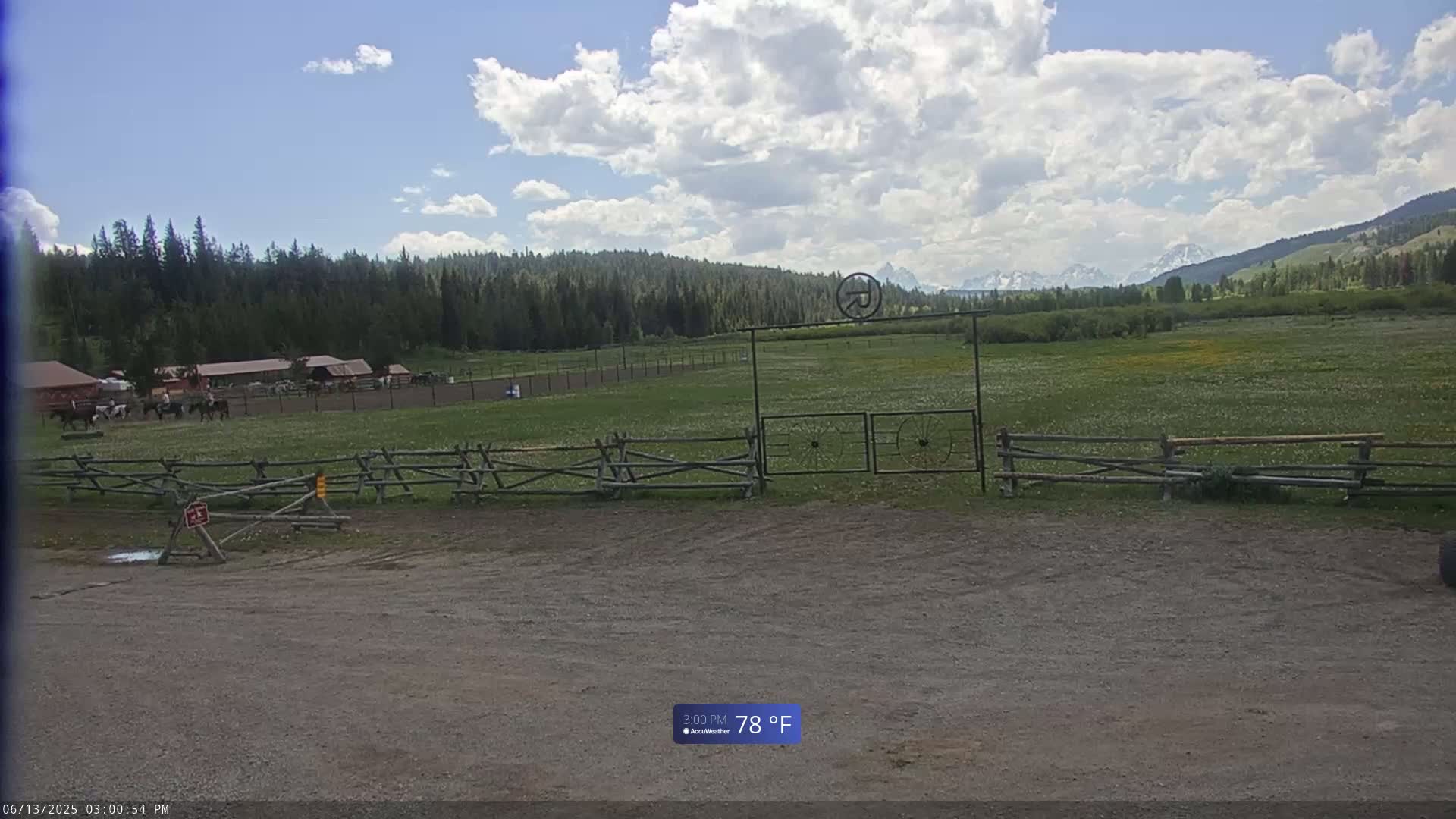 A grassy field with horses and a wooden fence sits in front of a mountainous background under a partly cloudy sky.