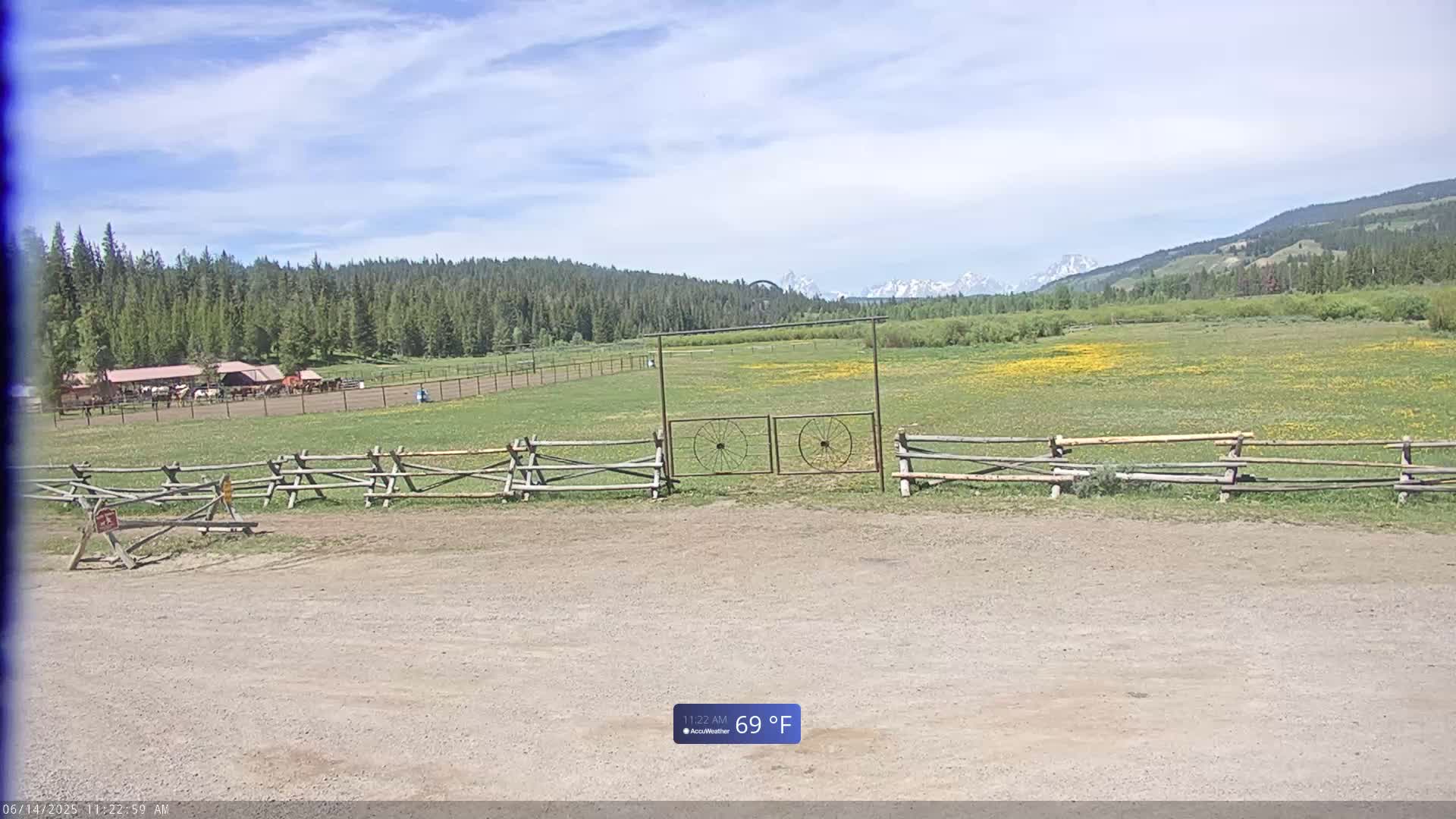 A dirt parking area is in front of a wooden split-rail fence and gate leading to a grassy field with wildflowers and a distant view of mountains under a partly cloudy sky.