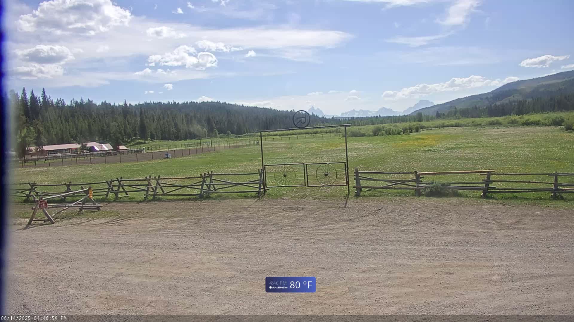 A dirt parking area is in front of a wooden split-rail fence and gate leading to a grassy field with wildflowers and a distant view of mountains under a partly cloudy sky.