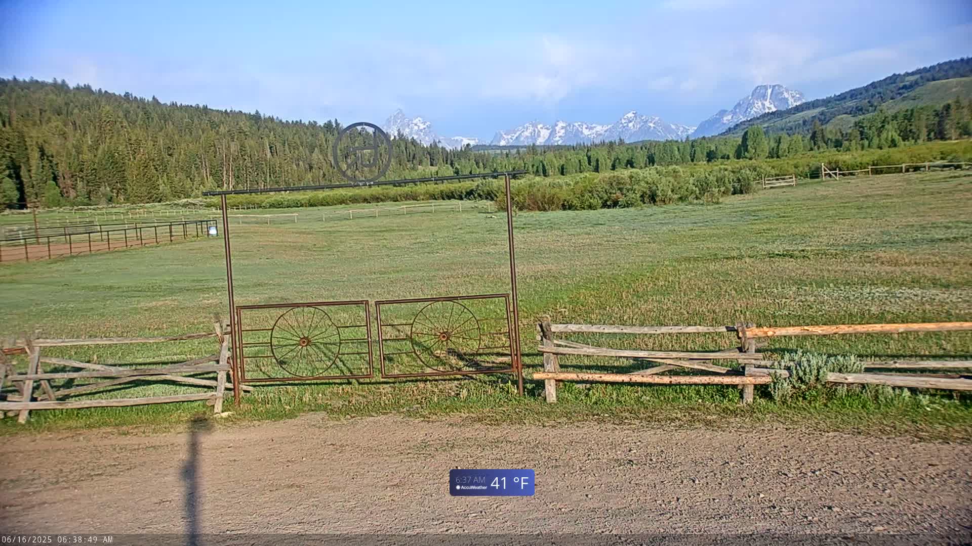 A grassy field with a split-rail fence and a metal gate featuring wagon wheels, backed by a forest and snow-capped mountains under a mostly clear sky.