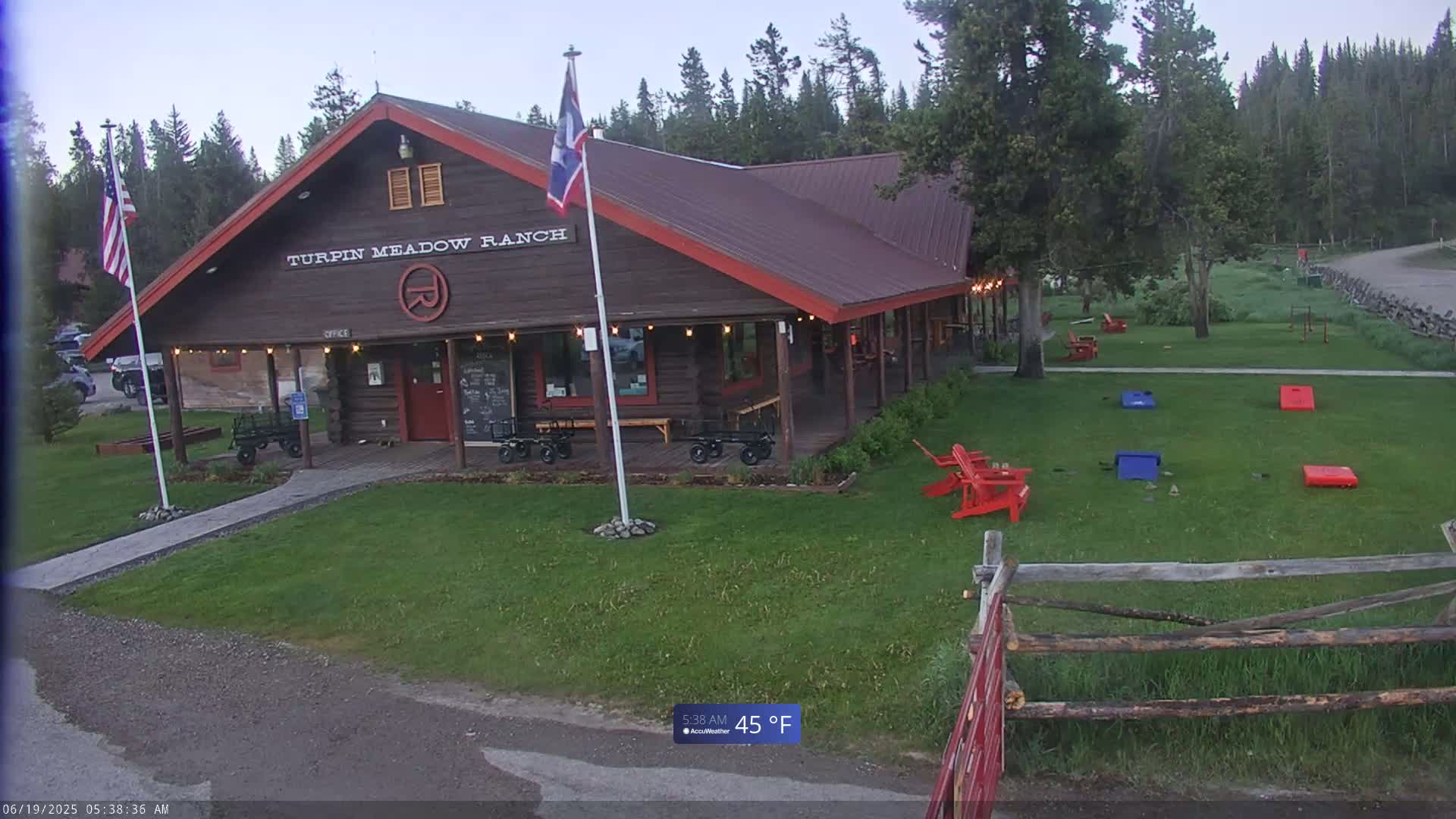 A log cabin building with a porch, American and another flag, and lawn games sits on a grassy area in front of a forest under a partly cloudy sky, with a temperature of 45°F.