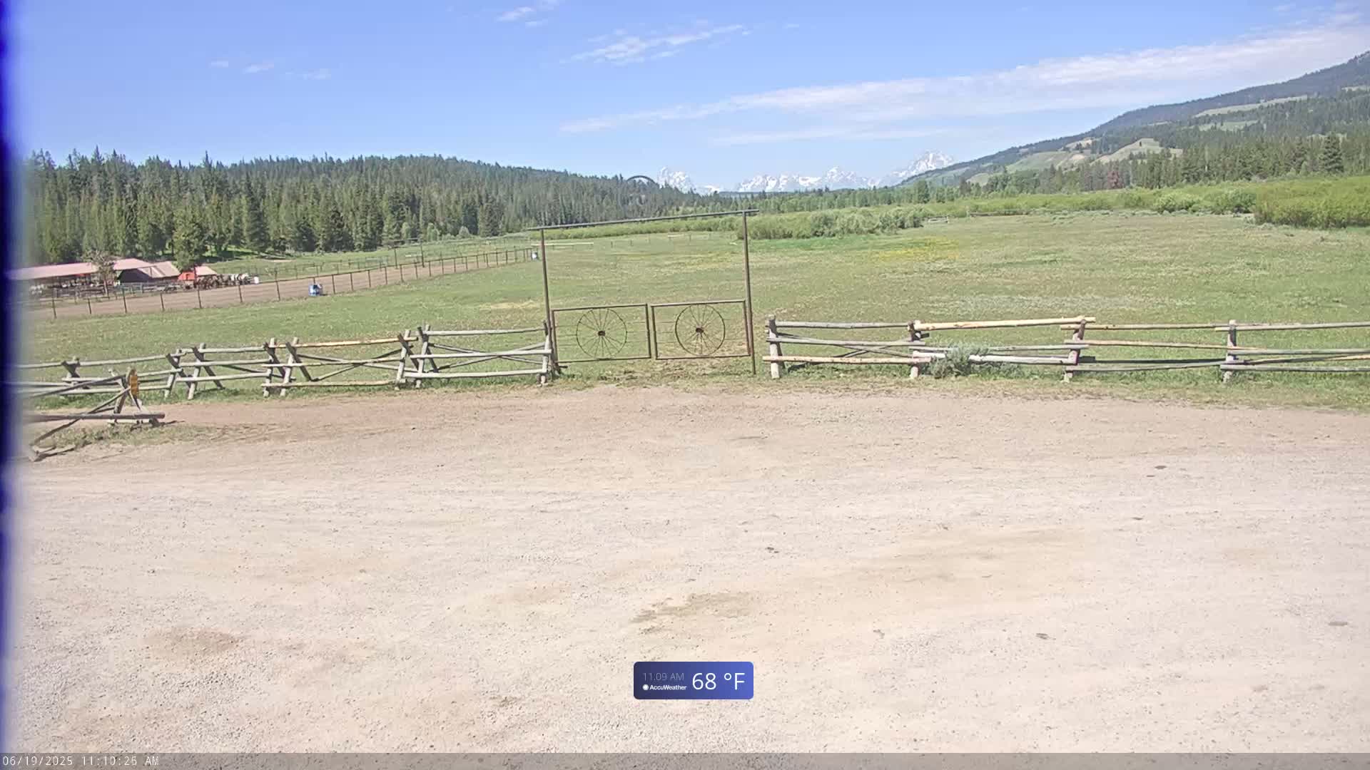 A dirt parking area is in front of a wooden fence and gate, beyond which is a grassy field, a line of trees, and snow-capped mountains under a clear blue sky.