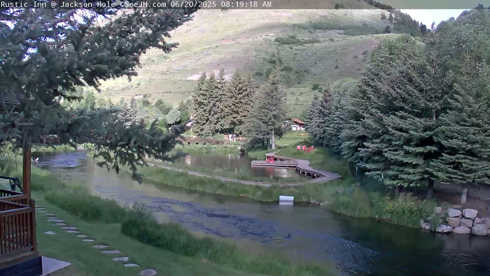 A tranquil river flows past a grassy area with a small pond and wooden dock, nestled amongst evergreen trees on a hillside under a partly sunny sky.