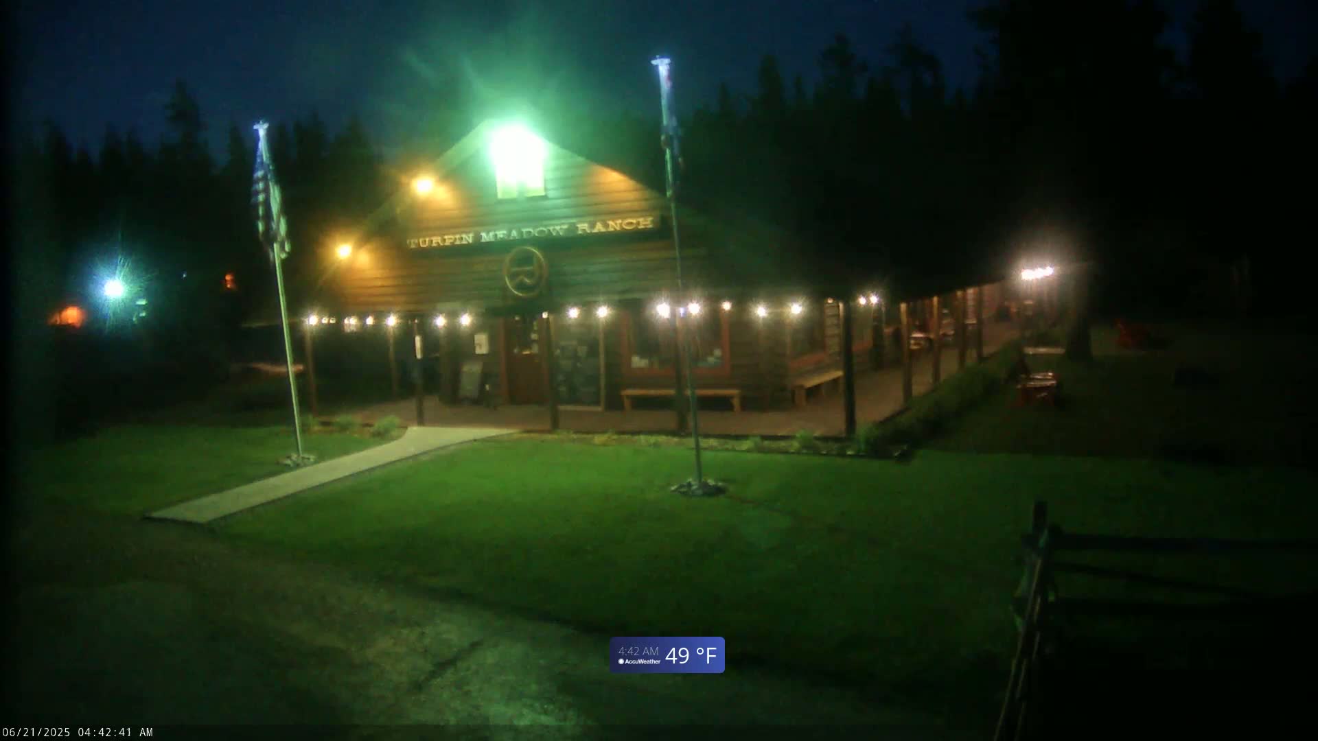 A lit-up wooden building with a porch sits on a grassy lawn at night under a dark sky; the temperature is 49°F.