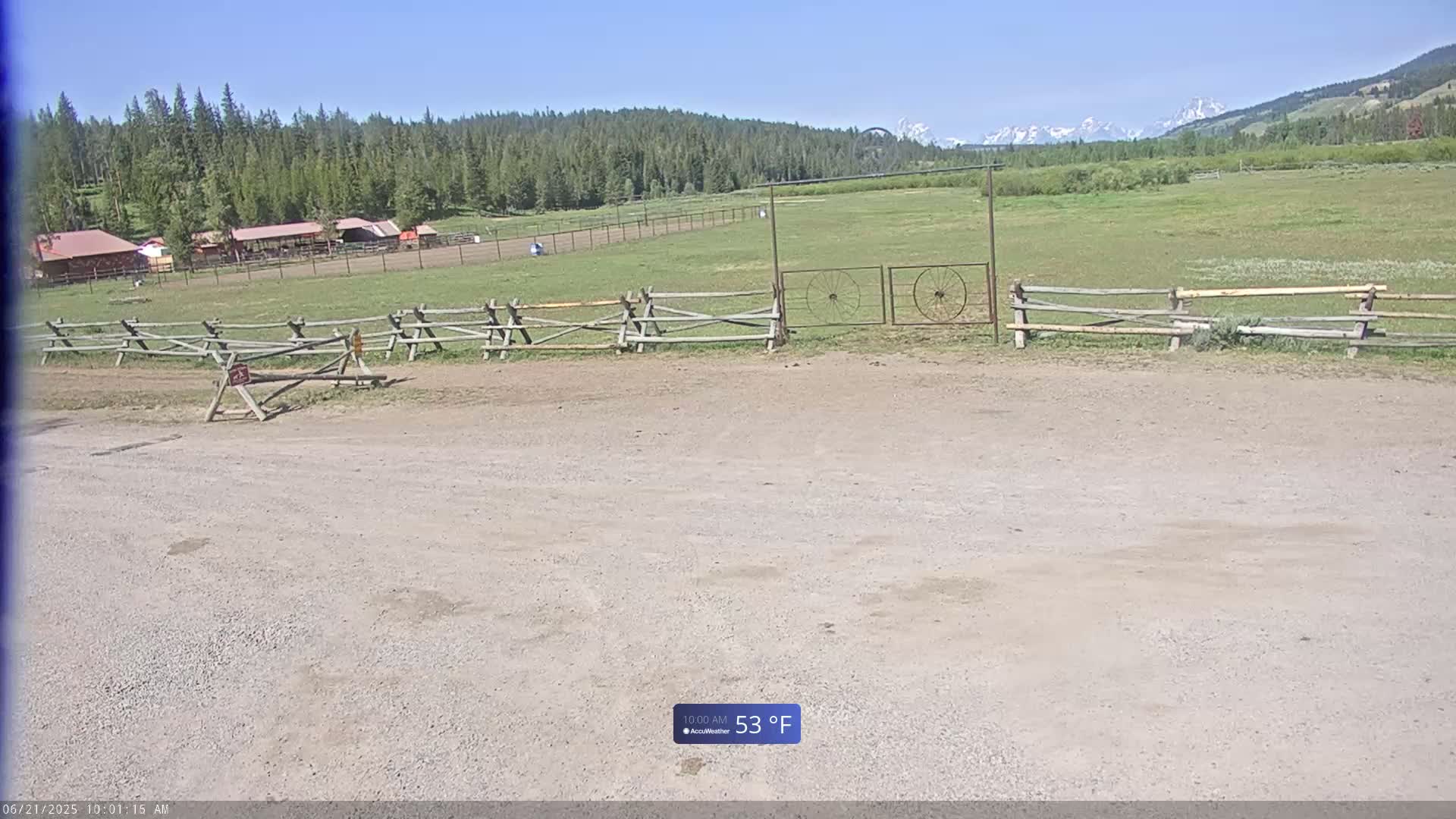 A gravel parking area sits before a wooden fence and gate, with a grassy field, distant buildings, a forest, and snow-capped mountains visible under a clear, sunny sky.