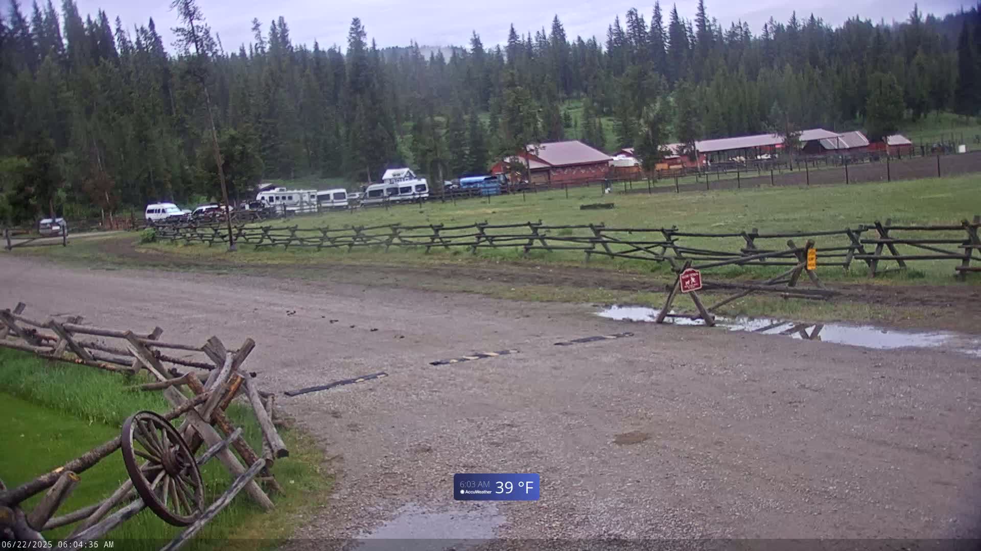 A gravel road leads to a building complex in a grassy field, bordered by wooden split-rail fences, with several vehicles parked nearby under overcast skies.