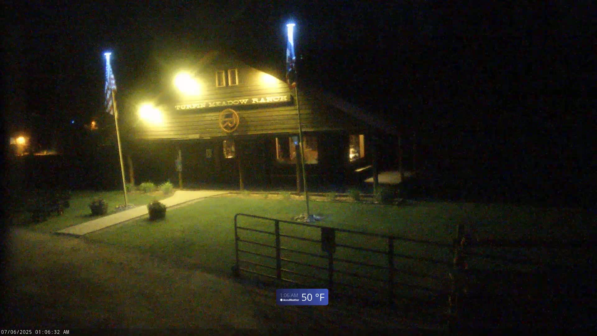 A dark-colored building with illuminated signage is visible at night, lit by artificial lights, with a fenced area and two flagpoles in front.