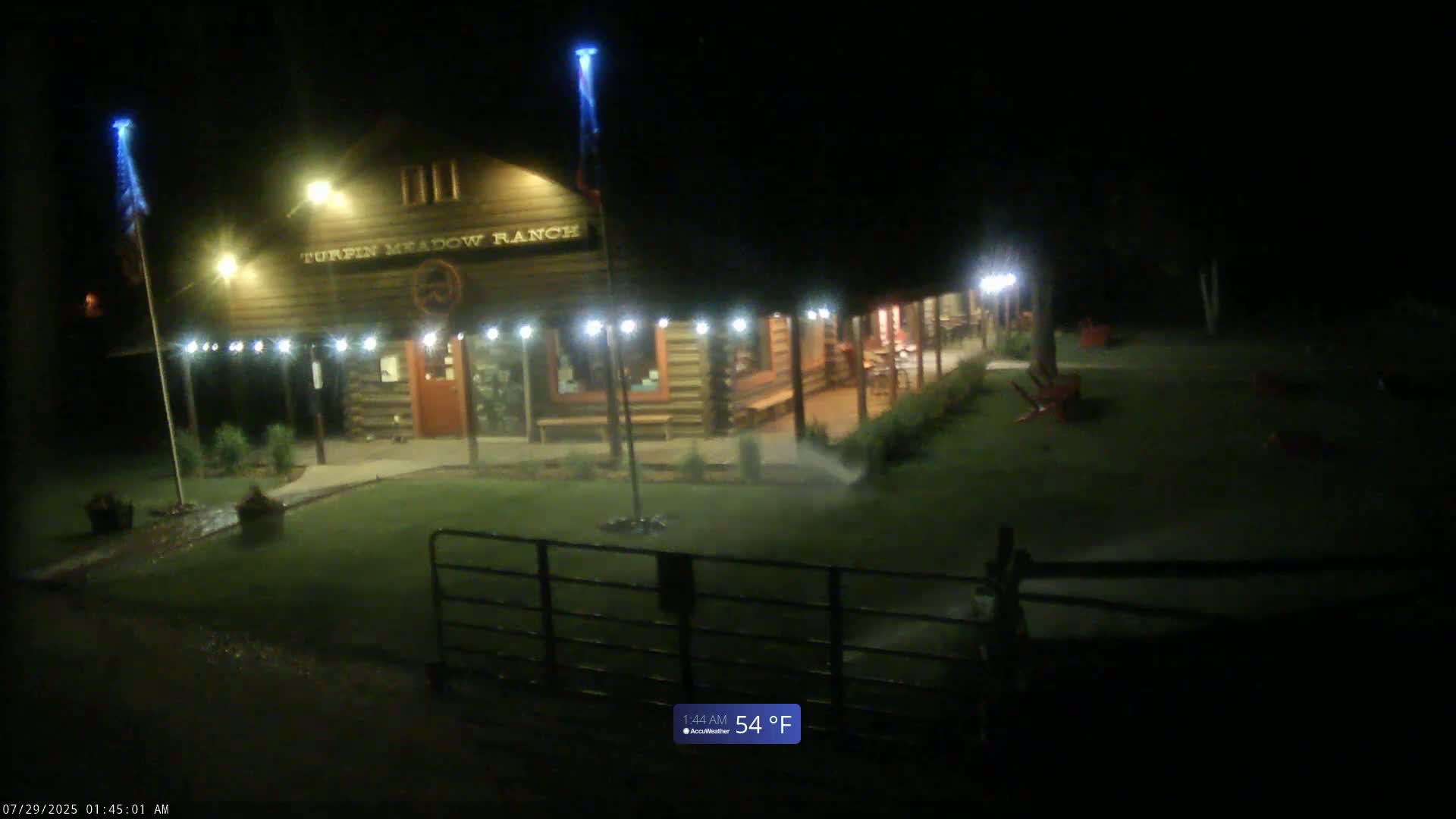 A lit-up log cabin building with a porch is being watered by sprinklers at night in 54°F weather.